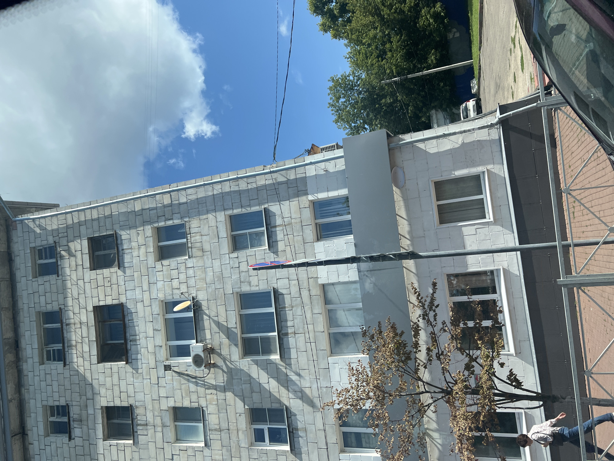 The image shows a modern apartment building with light gray, brick-like siding. The building has multiple floors with numerous windows, some of which have white frames. An air conditioning unit is visible mounted on the exterior wall of one of the lower floors. A small, sparsely leafed tree with brown foliage stands in front of the building. A woman in a patterned shirt and blue jeans is walking past the tree on a sidewalk, carrying a purse. She is walking from left to right. A metal fence with diagonal bars separates the sidewalk from the street. The sky is blue with scattered white clouds, and power lines are visible against the sky. In the background, there are green trees and a white car parked on a road. The overall scene appears to be an urban environment on a sunny day. The building's architecture and the visible signage (a red and blue circular sign on a pole) suggest a location in Russia. Specifically, the image was captured on Komsomol'skiy Prospekt in Ulyanovsk, Russia.