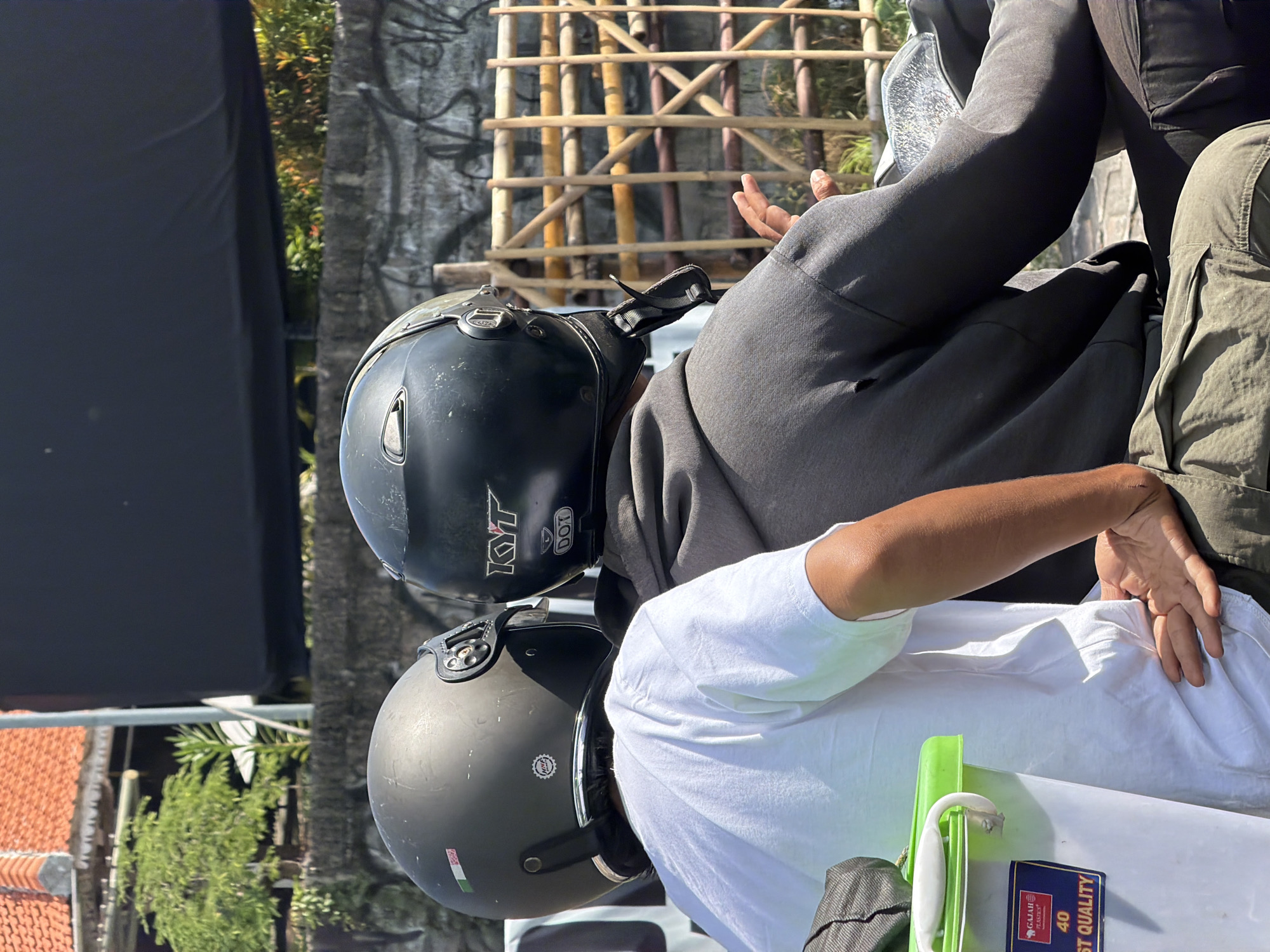 Two individuals wearing helmets are seated on a motorcycle. The person in the foreground wears a white t-shirt and is viewed from behind, with their left arm resting on their hip. The second individual, in the background, wears a grey hooded sweatshirt and a black helmet with "KYT" and "DOT" markings. They are also viewed from behind, with their right hand extended. The motorcycle is positioned outdoors, with a wall in the background featuring graffiti. A bamboo fence is visible behind the individuals. A white plastic bucket with a green lid and a label indicating "GAJAH PLASTICS" is situated to the left of the frame. The setting appears to be a street in Denpasar, Indonesia, under bright daylight.