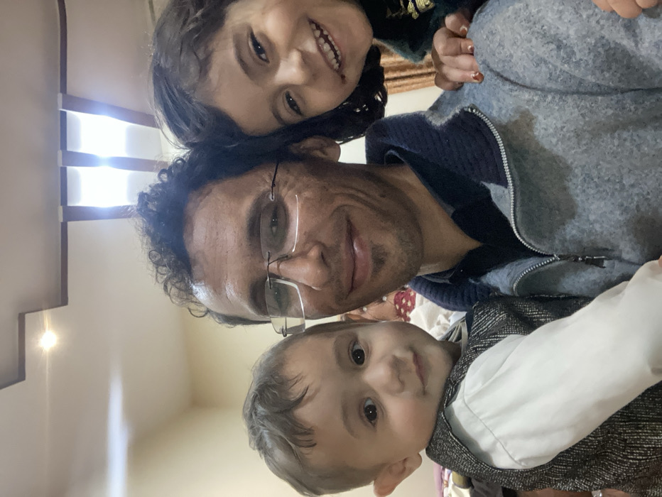 A man wearing glasses poses for a selfie with two children. The girl, positioned above the man, smiles broadly. The boy, below, looks directly at the camera. The man is wearing a zip-up sweater. The children's clothing details include a green top with gold accents and a white shirt with a patterned vest. The background shows part of a room with a light fixture. The image is rotated slightly counterclockwise.