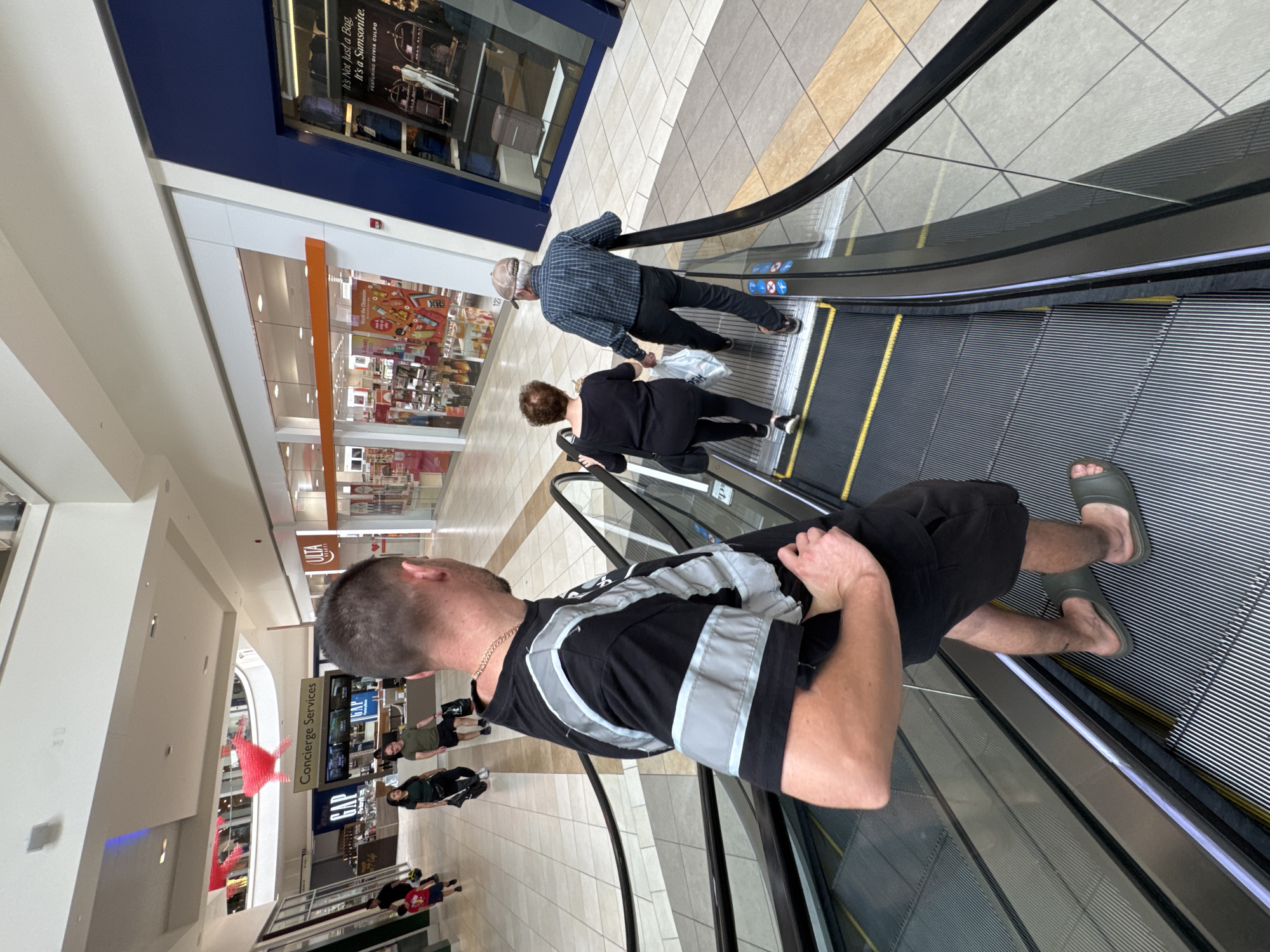 A group of people are on an escalator in a shopping mall. The man in the foreground is looking over his left shoulder, wearing a black t-shirt with reflective stripes, black shorts, and gray slide sandals. Behind him, an older couple is ascending the escalator; the woman is wearing a black shirt and pants, and the man is wearing a plaid shirt, dark pants, and a baseball cap. In the background, other shoppers are visible walking through the mall, and various store fronts, including "GAP" and "ULTA BEAUTY," are present. The mall appears to be well-lit with a tiled floor and a modern ceiling. The scene is taking place in Rosemont, United States of America.