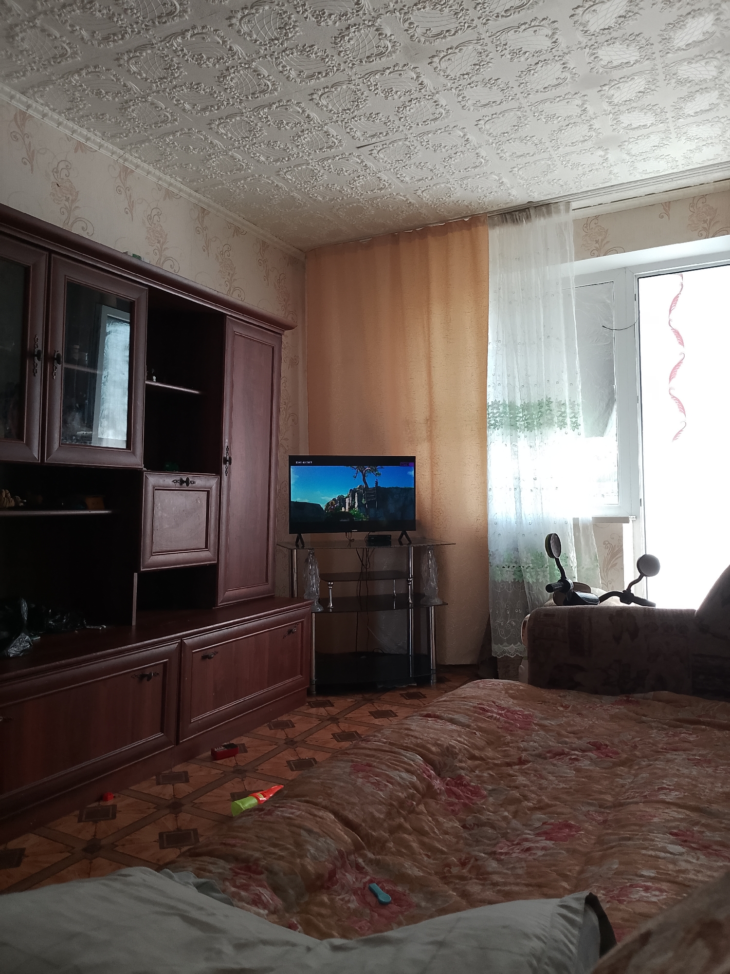 The image depicts an interior scene, likely a living room or bedroom, within a dwelling in the city of Abay, Kazakhstan. The room features a large, dark wood entertainment center with glass-fronted cabinets and shelves on the left side of the image. To the right of the entertainment center, a television is displayed on a black metal stand, showing a video game scene with a fantasy landscape.
The walls are covered with patterned wallpaper in a beige and floral design. The ceiling is decorated with embossed white tiles. A floor-to-ceiling window with sheer white curtains and an orange curtain is visible on the right side of the frame, allowing natural light to enter the room.
In the foreground, a large, floral-patterned blanket covers a bed or sofa. Various small objects, possibly toys, are scattered on the patterned floor, including a red rectangular object, a green and orange object, and a light blue object. A part of a gray pillow is visible in the lower left corner of the image, and a portion of a sofa with a patterned fabric is visible on the right. Two black handlebar-like objects, possibly from a toy or a piece of equipment, are situated near the window. The overall impression is of a lived-in domestic space.