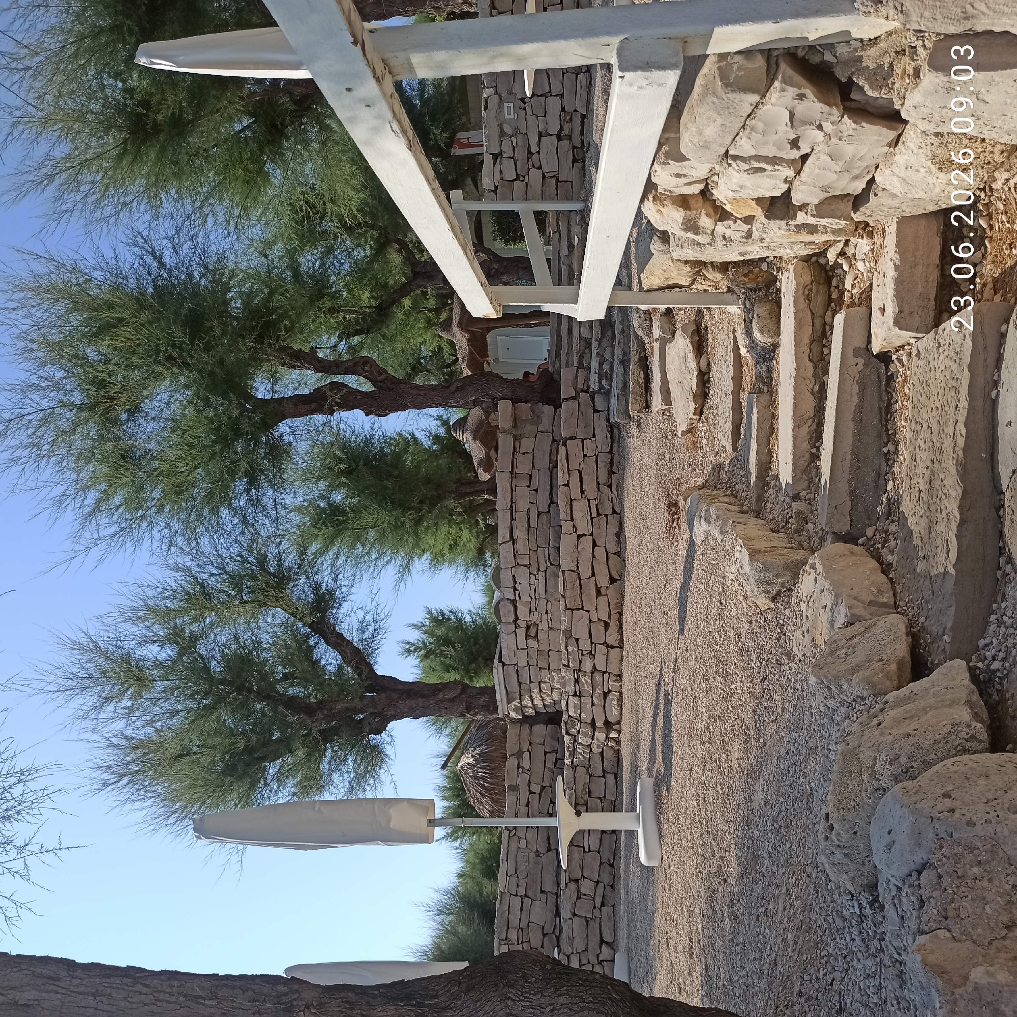 The image depicts an outdoor scene featuring a pathway leading up to a stone wall, with trees and foliage in the background. The pathway is made of uneven stone slabs and is bordered by large rocks and gravel. A white railing runs along the right side of the pathway. Behind the railing, a stone wall is visible, constructed from roughly hewn stones. In the background, several trees with sparse foliage are present, and beyond them, a structure with a thatched roof and a white door is partially visible. Two white, covered umbrellas or flags stand upright to the left of the pathway. The overall environment appears to be a well-lit, outdoor area with a natural, slightly rustic aesthetic. The image contains a timestamp in the lower right corner: "23.06.2026 09:03". There is a person seated on the steps to the right of the thatched roof structure. This location appears to be a beach or coastal area, possibly a resort or park, given the presence of umbrellas and the natural landscaping.