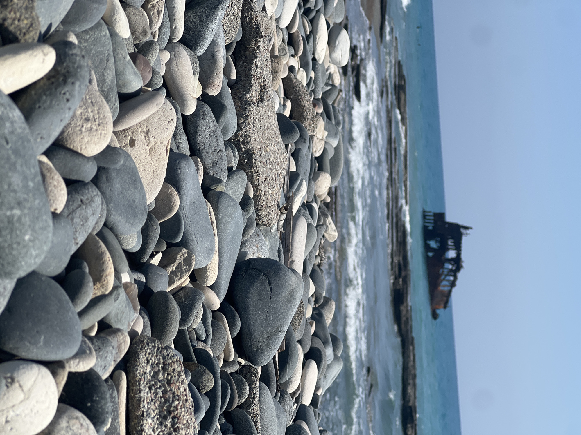 A shallow-angle, eye-level, medium shot captures a rocky beach with a rusted shipwreck visible in the background. The foreground is dominated by a dense collection of smooth, rounded pebbles in various shades of gray, white, and beige, forming a textured carpet that extends towards the sea. Some of these pebbles are large, with distinct shapes and surfaces. The midground shows a few larger, rougher rocks and some scattered debris, leading to the choppy, blue-green sea. Gentle waves roll in towards the shore, breaking against submerged rocky outcrops. In the distance, partially obscured by the waves and the shallow water, lies a decaying shipwreck. The ship appears to be a large vessel, heavily rusted and broken, with visible skeletal structures. The horizon is clear, meeting a pale blue sky. The overall scene depicts a weathered and rugged coastal environment, likely in Akrotiri, Sovereign Base Areas of Akrotiri and Dhekelia. There are no people visible in the frame.