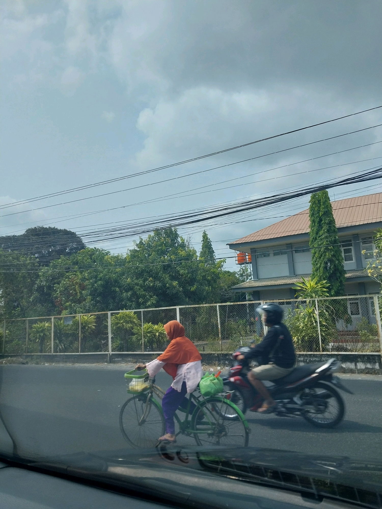A hazy, overcast sky looms above a street scene in Purwomartani, Indonesia. In the foreground, a person wearing an orange hijab and white and purple clothing rides a green bicycle with a green bag in the front basket. They are moving to the left. Behind them, a person in a black jacket and light-colored shorts rides a red and black motorcycle to the left, wearing a black helmet. The road is paved and appears wet or reflective. Beyond the road, a chain-link fence borders a green, manicured area with various plants and trees. In the background, a two-story building with a brown roof is visible, alongside more trees and utility wires crisscrossing the sky. A large orange water tank sits atop a metal structure to the left of the building. The overall impression is one of an everyday scene on a cloudy day in a semi-urban environment.