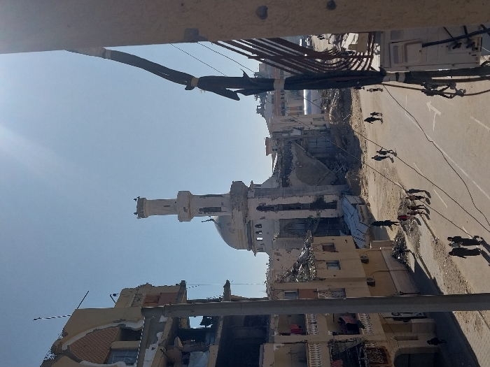 The image depicts a severely damaged urban area in Khan Yunis, Palestinian Territory, under a bright, clear sky. In the background, a large mosque with a white dome and a tall, slender minaret stands prominently; the main structure of the mosque behind the minaret is heavily damaged, exhibiting collapsed walls and exposed interior elements. To the left, a multi-story building shows significant structural compromise, with upper levels partially demolished, exposed rebar, and laundry hanging from lower, partially intact sections. A paved road, marked with white directional arrows, traverses the scene. Approximately 15 individuals, comprising adults and children, are observed walking on the road and sidewalks. In the foreground-left, a group of women, some wearing head coverings, walk together. The immediate right foreground shows the side of a building with multiple dark cables running vertically and horizontally. The ground around the structures is covered in dirt and rubble, consistent with extensive destruction.