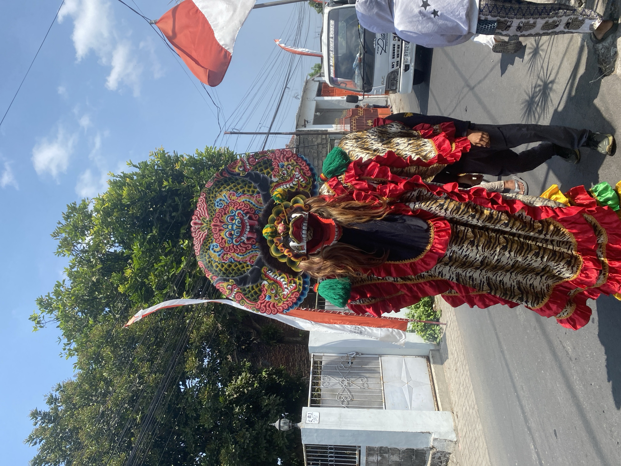 A person in an elaborate, colorful costume resembling a mythical creature walks down a street in Kediri City, Indonesia. The costume features a large, detailed mask with a fierce expression, long brown hair, and a headdress adorned with intricate patterns. The body of the costume is draped with red ruffles and a tiger-striped fabric. Two Indonesian flags, red and white, are visible in the background, one on a pole and another on a vehicle. A white truck with the license plate "AG 8702 E" is parked on the side of the road. Other people are also present, some walking alongside the costumed individual and others observing from the side. The scene appears to be a celebration or parade. The environment is a street with houses and trees lining the sides, under a clear blue sky with some clouds.