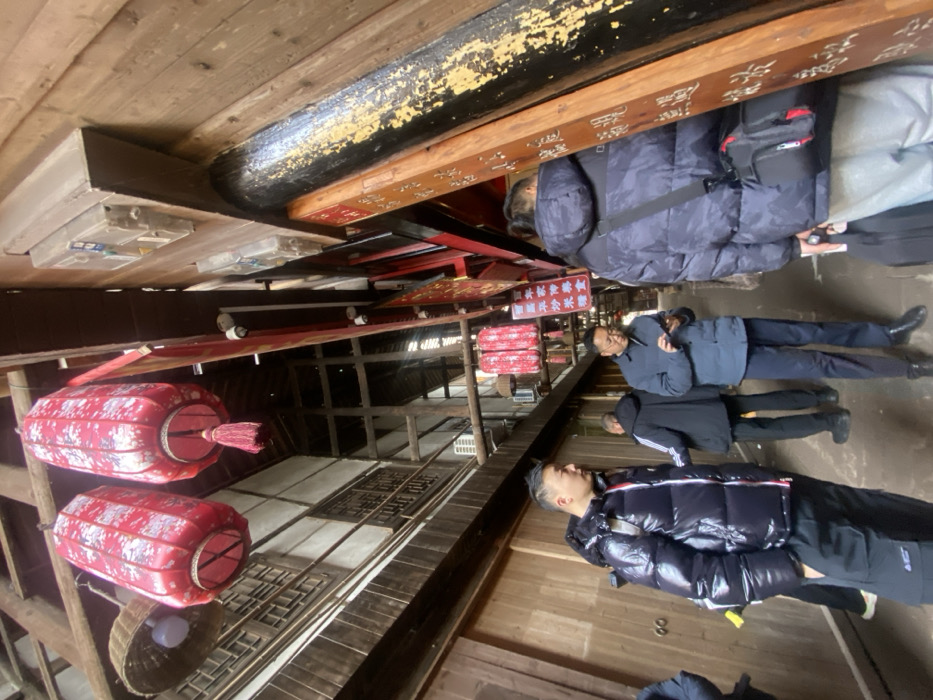 The photo captures an indoor scene, likely a street in a historical district. Red lanterns hang from the ceiling above wooden structures. Several people are seen walking through the area, with some wearing puffy jackets. There are signs with Chinese characters displayed. The space has an old, traditional atmosphere.