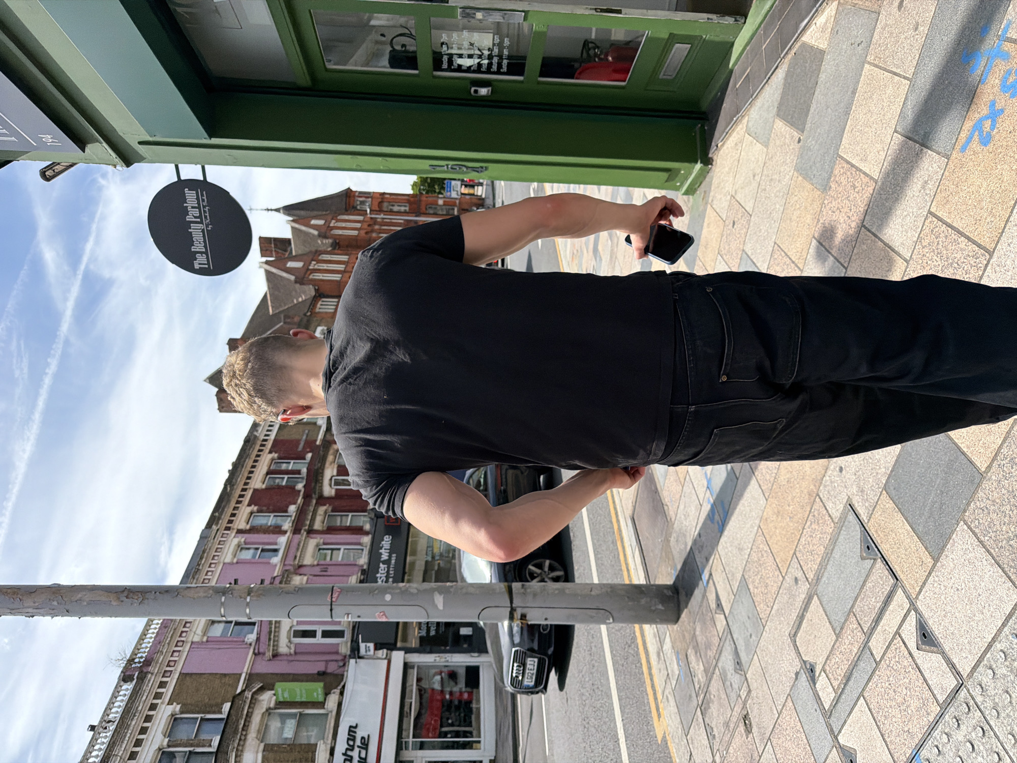 A man with blonde hair is walking away from the camera on a sidewalk in London, United Kingdom. He is wearing a black t-shirt and dark jeans, and he is holding a black smartphone in his right hand. The sidewalk is paved with multicolored square tiles, and there are several metal utility covers. A gray metal pole stands to the left of the man. In the background, there are several buildings, including a pink building with white trim and windows, and a green building with a sign that reads "The Beauty Parlour". An Audi car is parked on the street in front of the pink building. The sky is blue with white clouds.
