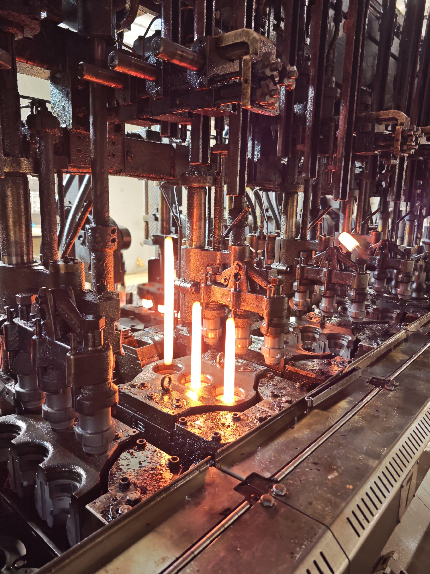 The image shows an industrial setting, likely a glass-making factory, with multiple mechanical arms and components coated in a dark substance, possibly oil or grease. At least three molten glass rods are visible, glowing with intense heat. The machinery is intricate and appears to be part of an automated production line. A metal conveyor belt runs along the bottom right of the image. The location is not discernible from the image alone, and I am unable to determine if it is in Pleven, Bulgaria.