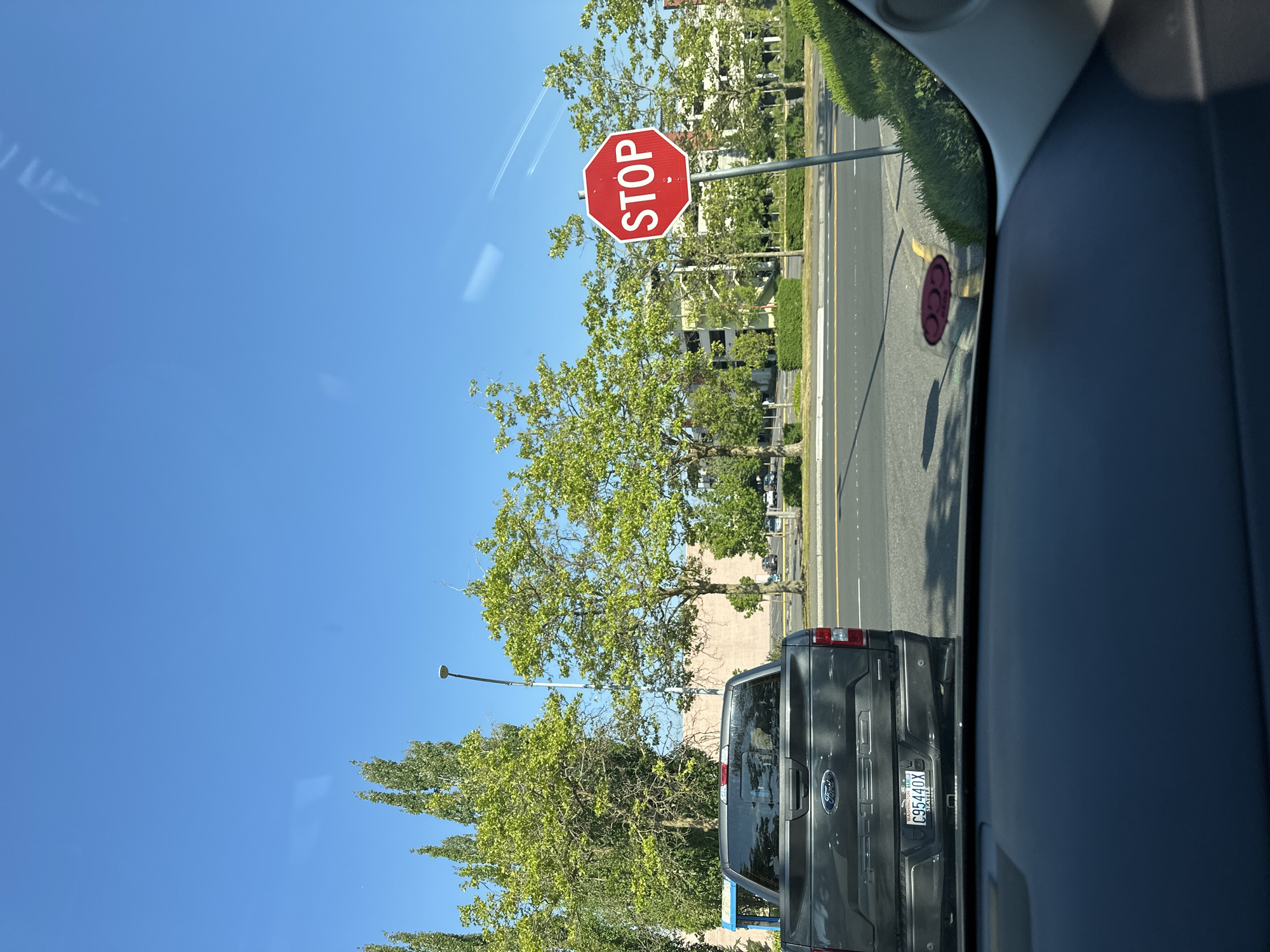 The image depicts a view from inside a vehicle looking towards a street with a stop sign. A dark grey Ford F-150 pickup truck is parked on the right side of the road, facing away from the camera. The truck has a Washington State license plate that reads "C95440X SEATTLE". To the right of the truck, a red octagonal stop sign is prominently displayed on a metal pole. Beyond the stop sign, a multi-story building, possibly a parking garage or office building, can be seen. Lush green trees line the street, and the sky is a clear, bright blue with faint contrails from an aircraft. The foreground shows the dashboard and windshield of the vehicle from which the photo was taken, with reflections visible on the glass. The scene appears to be located in a commercial or suburban area of Lynnwood, United States of America.