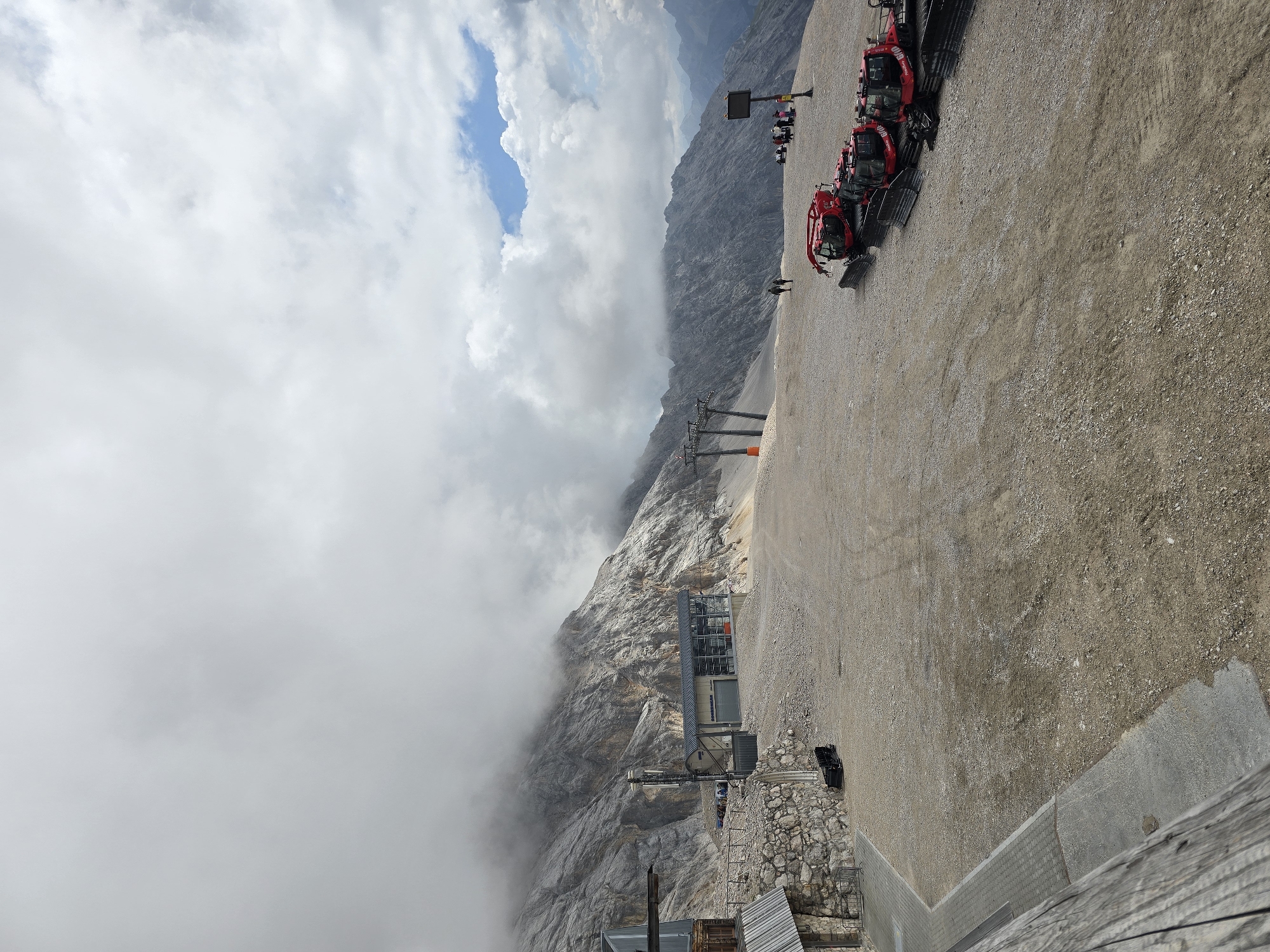 This image depicts a mountain landscape near the Zugspitze cable car station, in Garmisch-Partenkirchen, Germany. The foreground is a gravelly, rocky area, with a pathway made of concrete and paving stones on the left. Several red snow groomer vehicles are parked in a line on the right side of the image. In the midground, a modern building with large glass windows, labeled "SONNENKAR", is situated at the base of a steep, rocky mountain. A cable car pylon is visible to the left of this building, and another pylon is seen further in the distance. The mountain slopes are covered in rock and some sparse vegetation, with low-lying clouds obscuring the upper reaches of the mountains. In the far distance, a range of further mountains is visible under a partly cloudy sky with patches of blue. A few individuals are walking in the midground, and a small group of people are gathered near a signpost in the right midground. The overall scene conveys an alpine environment, likely a tourist area, with a focus on infrastructure for mountain transport and maintenance.