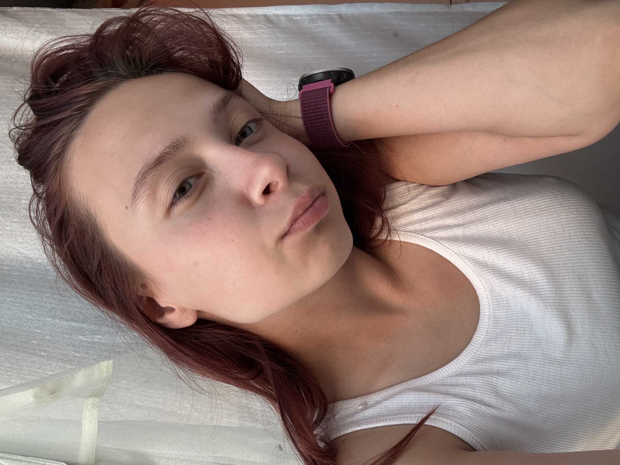 A young Caucasian woman with auburn hair is captured in a close-up, selfie-style shot. She is wearing a white ribbed tank top and has her left arm bent, supporting her head with her hand. A purple watch with a pink accent is visible on her left wrist. The background consists of white and light blue sheer curtains. The lighting suggests an indoor setting with natural light filtering through the window. No specific location in Прикамский, Russia, can be determined from the image.