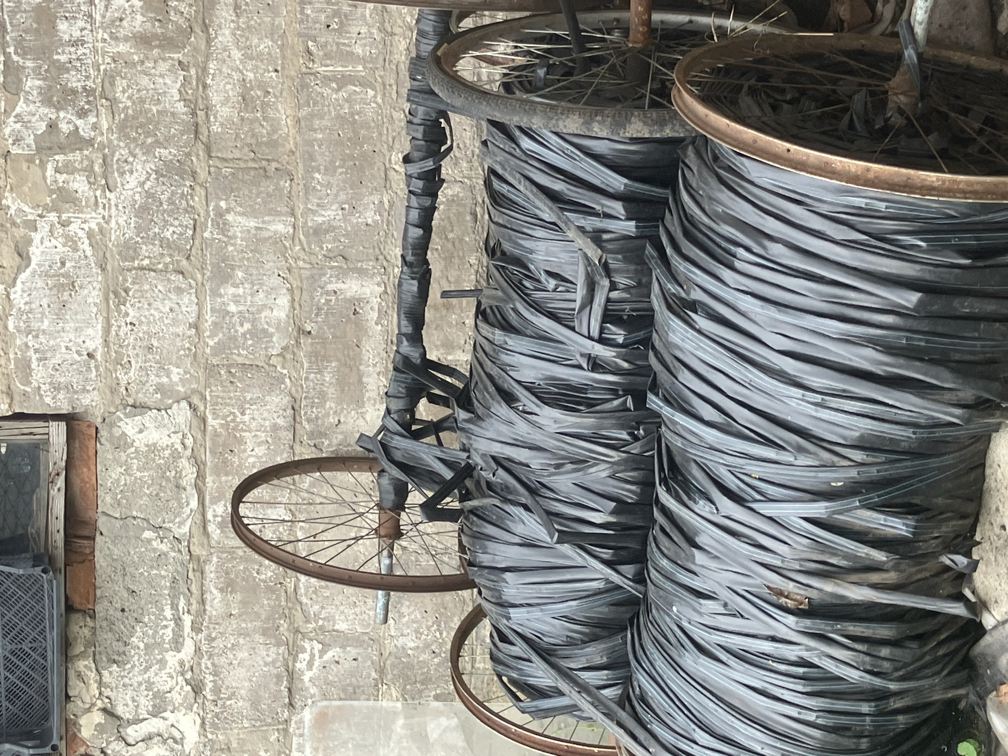 In Vovkivka, Ukraine, two large rolls of black, plastic-like material are stacked on top of each other, supported by metal frames with spoked wheels. The material appears to be a continuous, wide strip wound into dense cylindrical forms. Above the rolls, a metal bar wrapped in similar black material is suspended, also supported by a wheel. In the background, a textured, weathered brick wall serves as the backdrop, with a section of a grey, slatted object visible near the top. The ground beneath the rolls is rough and appears to be a mix of dirt and debris. The overall scene suggests an outdoor storage or work area with discarded or repurposed industrial materials.