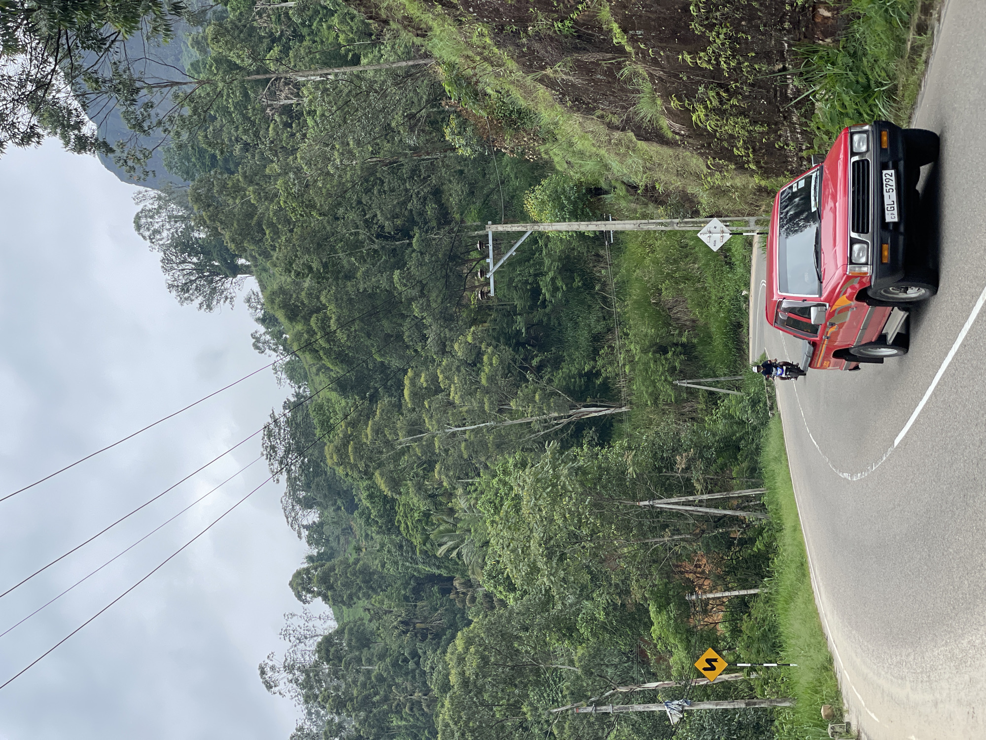 The image depicts a winding road in Tawalantenna, Sri Lanka, surrounded by dense vegetation. A red pickup truck with license plate "GL-5792" is on the road, and a motorcyclist is visible next to the truck. Utility poles and wires run along the road. A yellow "S" curve road sign is located near the road. The environment is lush and hilly.