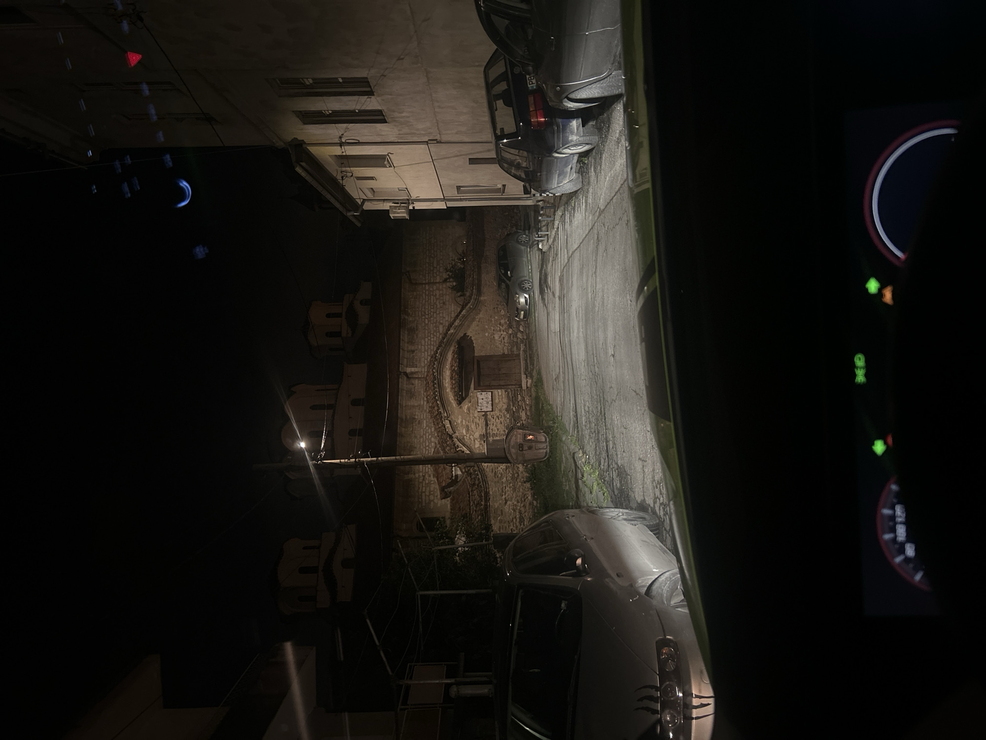 Captured from inside a vehicle, the image reveals a night view in what seems to be an urban alleyway within the city of Unknown, Unknown. Cars line the right side of the alley, while the left shows the dark silhouettes of buildings. A streetlight illuminates the alley, casting light on the cobblestone paving, and a brick wall structure is visible further down the alley. The car's dashboard is visible on the right of the image, with the speedometer and possibly other indicators lit. The overall setting is dimly lit, adding to the mysterious feel of the urban environment.