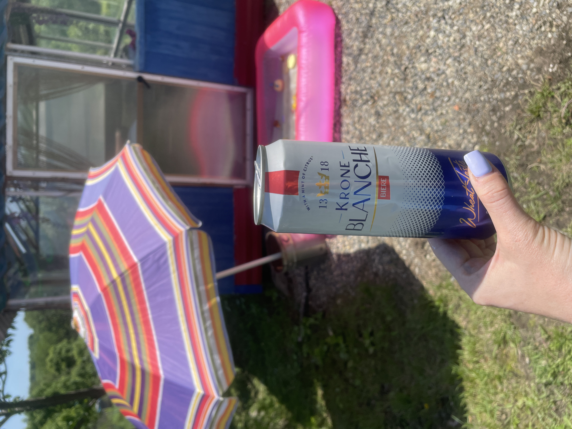 A person's hand with light purple fingernail polish is holding a can of Krone Blanche Biere in an outdoor setting. The can has a white and blue label with gold lettering. Behind the can, a colorful striped umbrella is partially visible, casting a shadow on the ground. In the background, there is a small inflatable pink pool with some yellow objects inside. Further back, a blue structure with a glass door is visible. The environment appears to be a sunny day with green grass and trees. The location is identified as the city of Yasnoe, Russia.