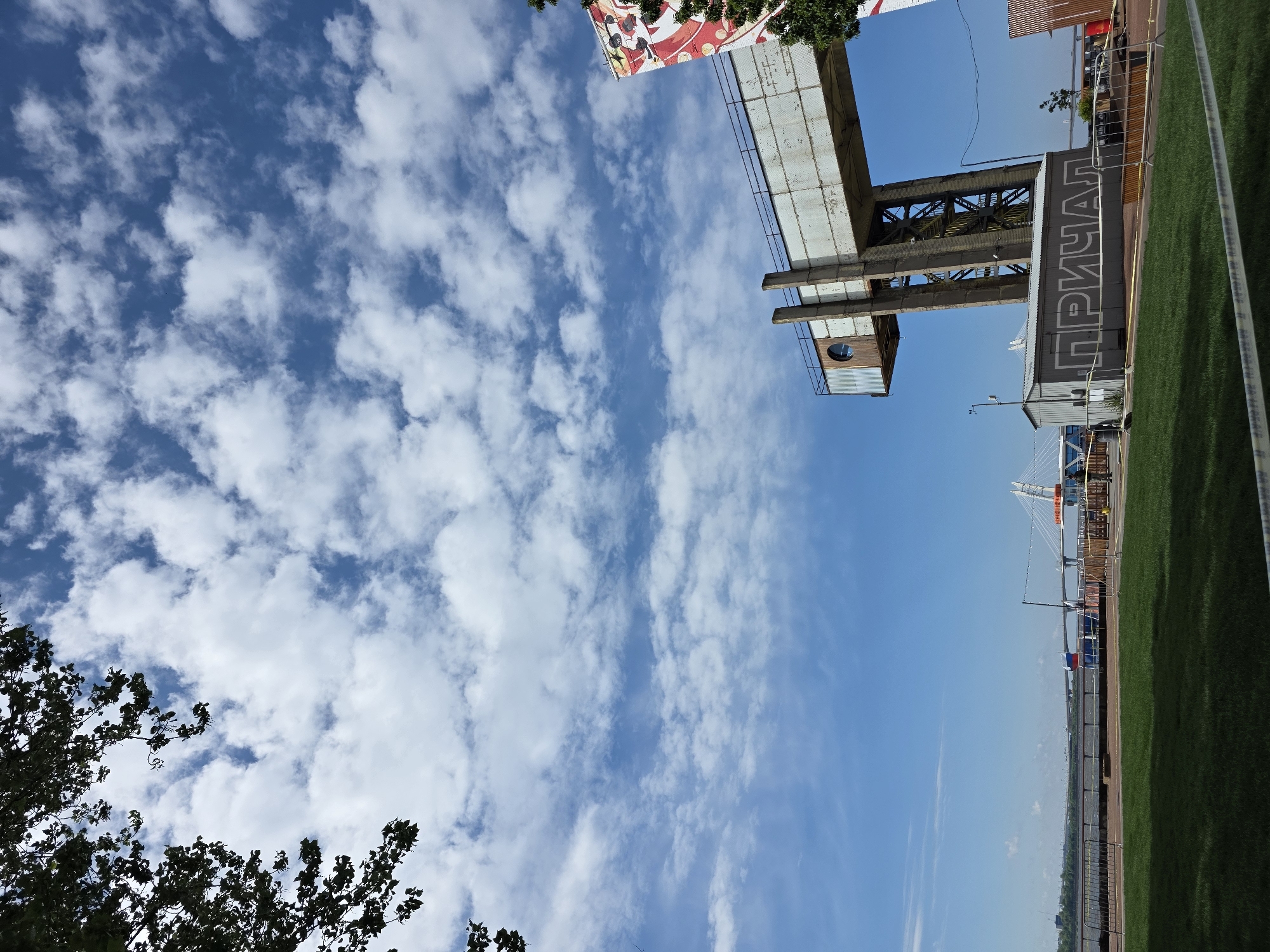 The image shows a sideways view of a landscape featuring a structure with Cyrillic text, open blue sky with scattered white clouds, and partial views of trees and greenery. The structure with Cyrillic text, reading "ПРИЧАЛ", appears to be part of a pier or docking facility (prichal meaning pier or dock). A nearby bridge, most likely the Saint Petersburg Stadium Bridge, is seen in the distance. The scene is likely in Saint Petersburg, Russia, given the Cyrillic text and architecture.
