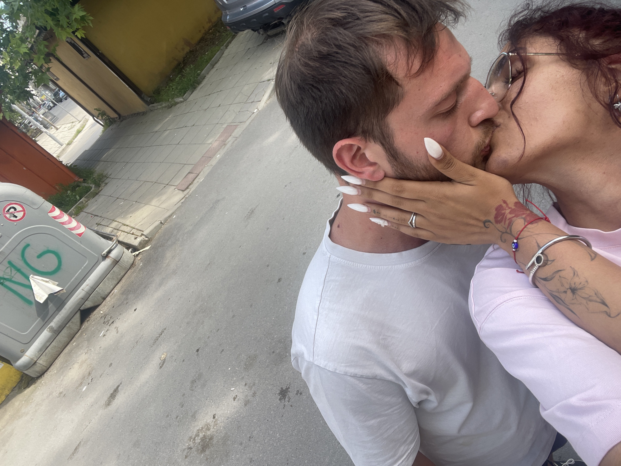 An overhead, slightly tilted photograph captures two individuals, a male and a female, kissing on a street in an urban environment located in Unknown, Unknown. The male, positioned on the left, wears a white t-shirt and has short brown hair and facial hair. The female, on the right, wears a light pink top, has dark, curly hair, and wears glasses. Her right hand, adorned with long white pointed nails, a silver ring, a silver bangle, and a red string bracelet with a blue 'evil eye' bead, is placed on the male's neck/jawline. Tattoos are visible on her right forearm and wrist, including floral designs. Both individuals have their eyes closed during the kiss. The background features a grey asphalt road with stains, a tiled sidewalk, and various structures. A large grey rectangular trash can with green "NG" graffiti and red and white striped reflective tape is prominent in the upper left. A partial red and white circular sign is also visible near the trash can. Further in the background, a yellow building, a reddish-brown wall, green foliage, and parked cars are discernible. The scene is illuminated by bright daylight.