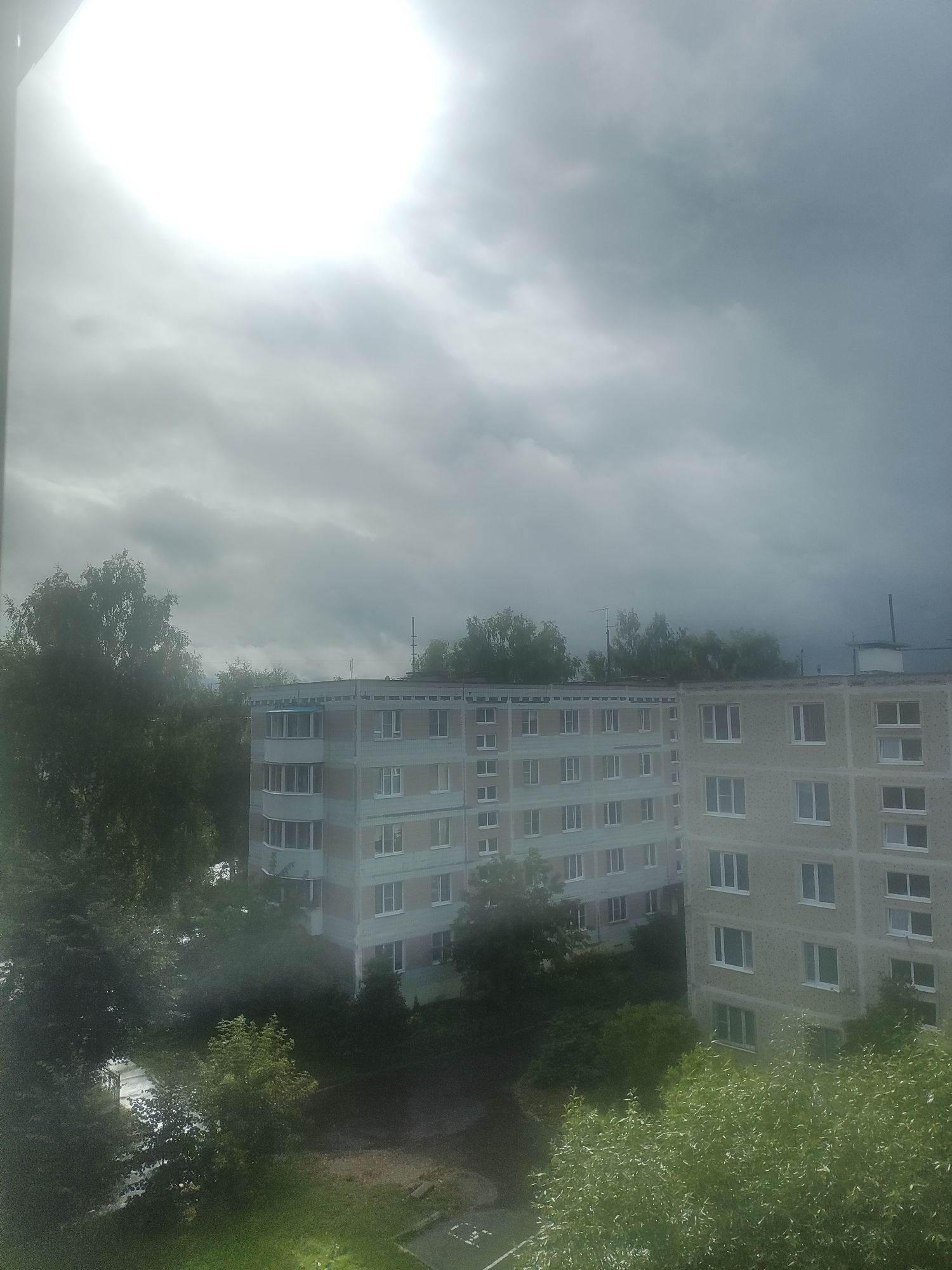 The image captures a view from a window in пгт Вербилки, Russia, overlooking two adjacent apartment buildings under a cloudy sky with the sun partially obscured. The apartment buildings appear to be of a Soviet-era design, with multiple floors and numerous windows. Trees and greenery surround the buildings and are visible in the foreground and between them. The lighting suggests it is daytime, but the cloudy conditions create a diffused and somewhat dim atmosphere. No people or specific actions are discernible in the scene.
