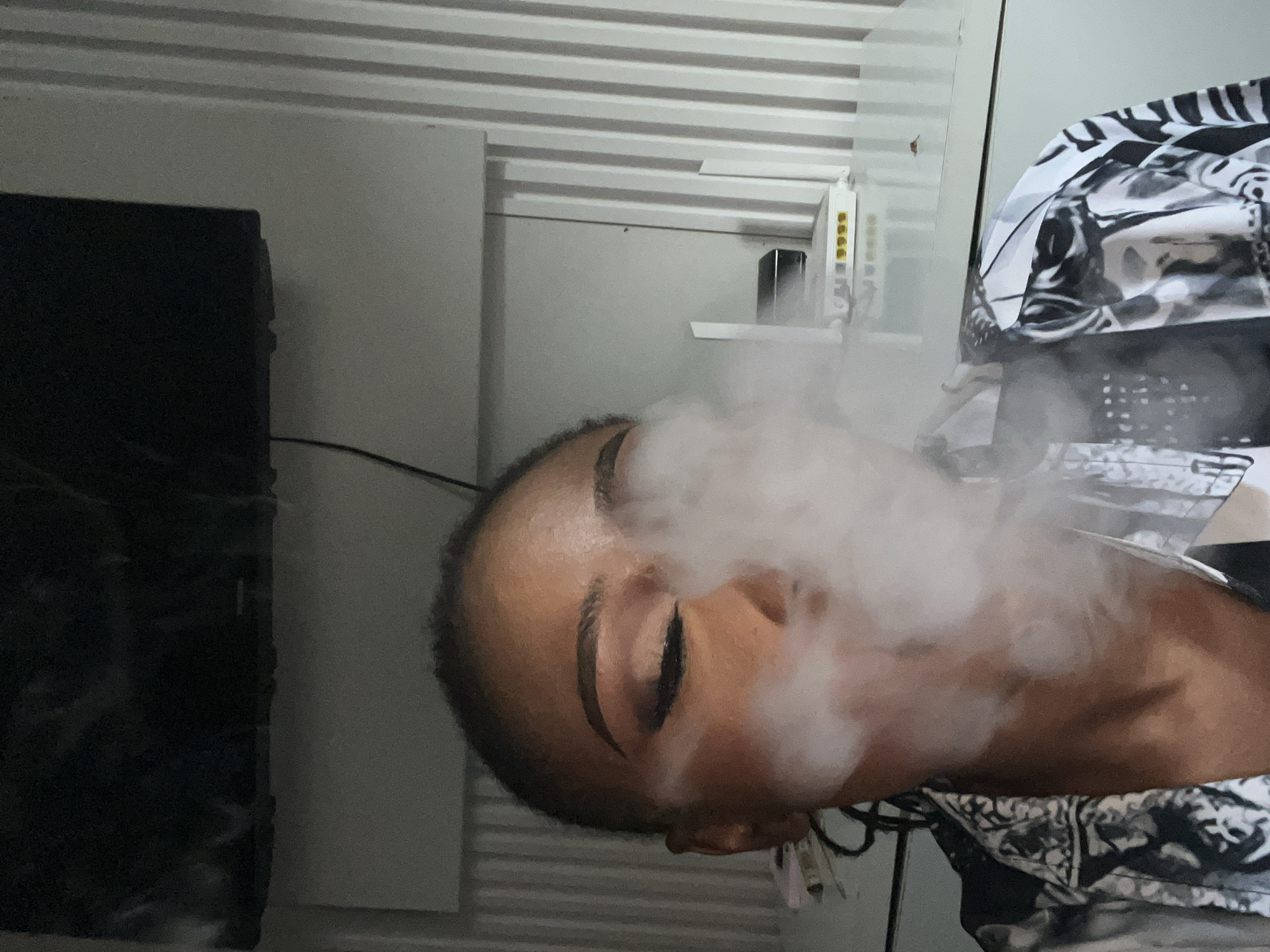 In an indoor setting, a person exhales a cloud of smoke, obscuring their face. The individual is wearing a black and white patterned shirt. Behind them, a white router with yellow ports sits on a surface, and a fluted wall forms the background. The scene appears to be in a residential area within Abuja, Nigeria, possibly a bedroom or living room.