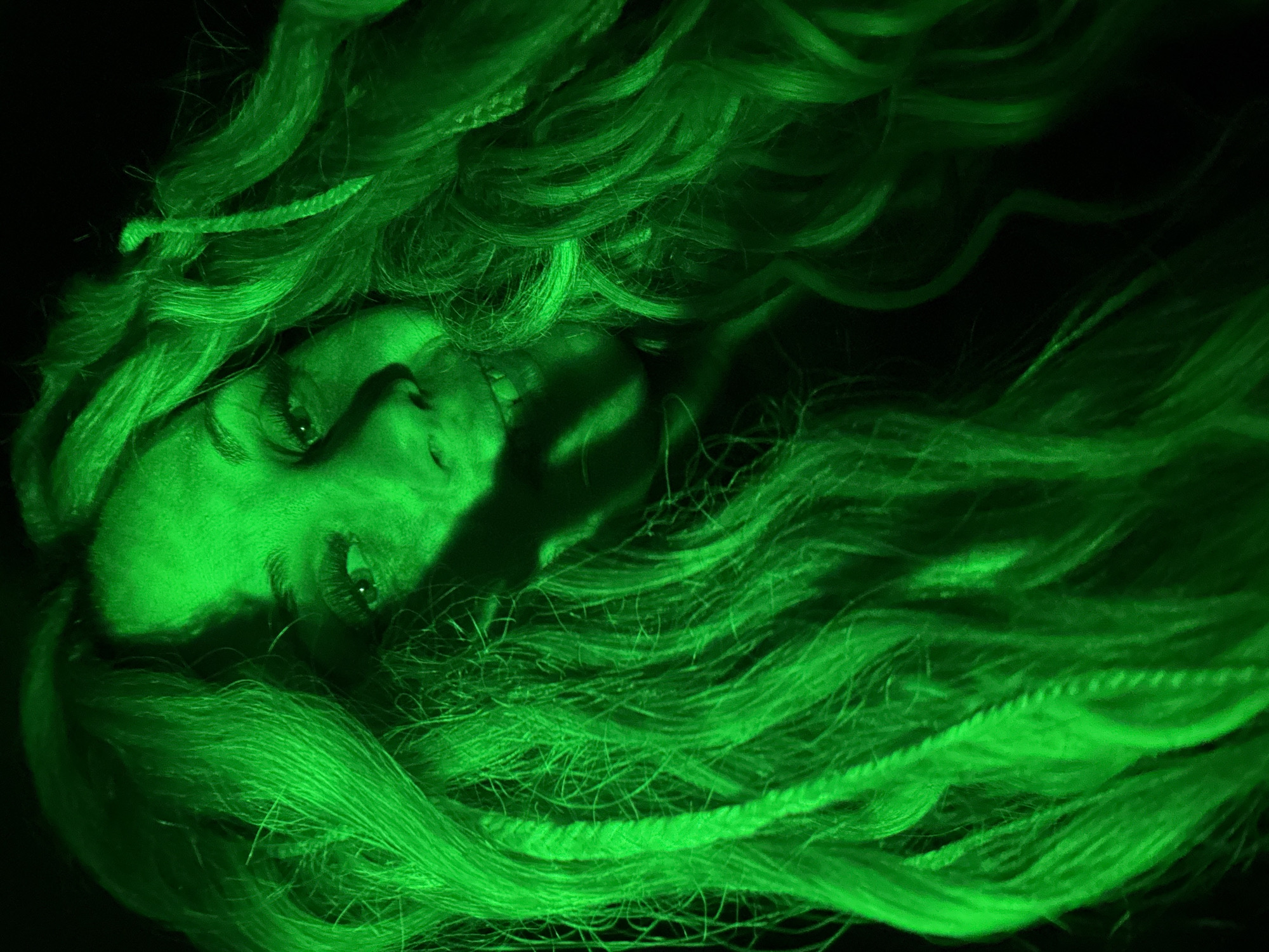 The image depicts a person with long, flowing green hair against a dark background. The person's face is partially visible, with their eyes open and looking towards the viewer. The overall scene is bathed in a green light, possibly due to artificial lighting or post-processing. There is no information in the image to support the claim that the photo was taken in Rusanovo, Russia.