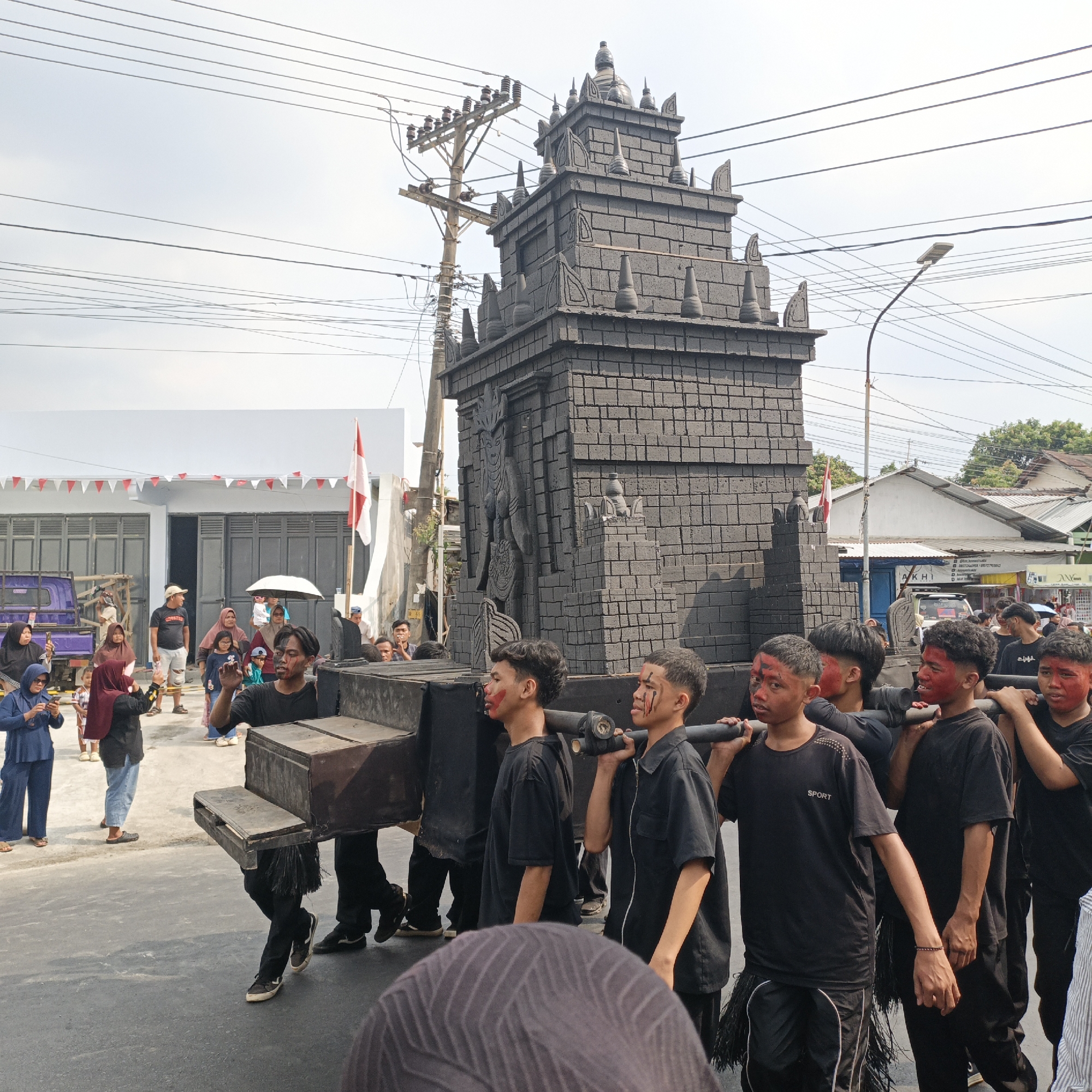 In Randudongkal, Indonesia, a group of young men dressed in black, with red paint on their faces, are carrying a large, dark, brick-like structure on their shoulders. The structure resembles a stylized, multi-tiered temple or monument with decorative spires and a carved relief on its facade. They are walking on a paved street, with onlookers gathered to the sides. A purple truck is visible in the background, along with buildings adorned with red and white triangular flags. Power lines crisscross the sky above, and a street lamp stands to the right. The scene suggests a procession or cultural event taking place outdoors.