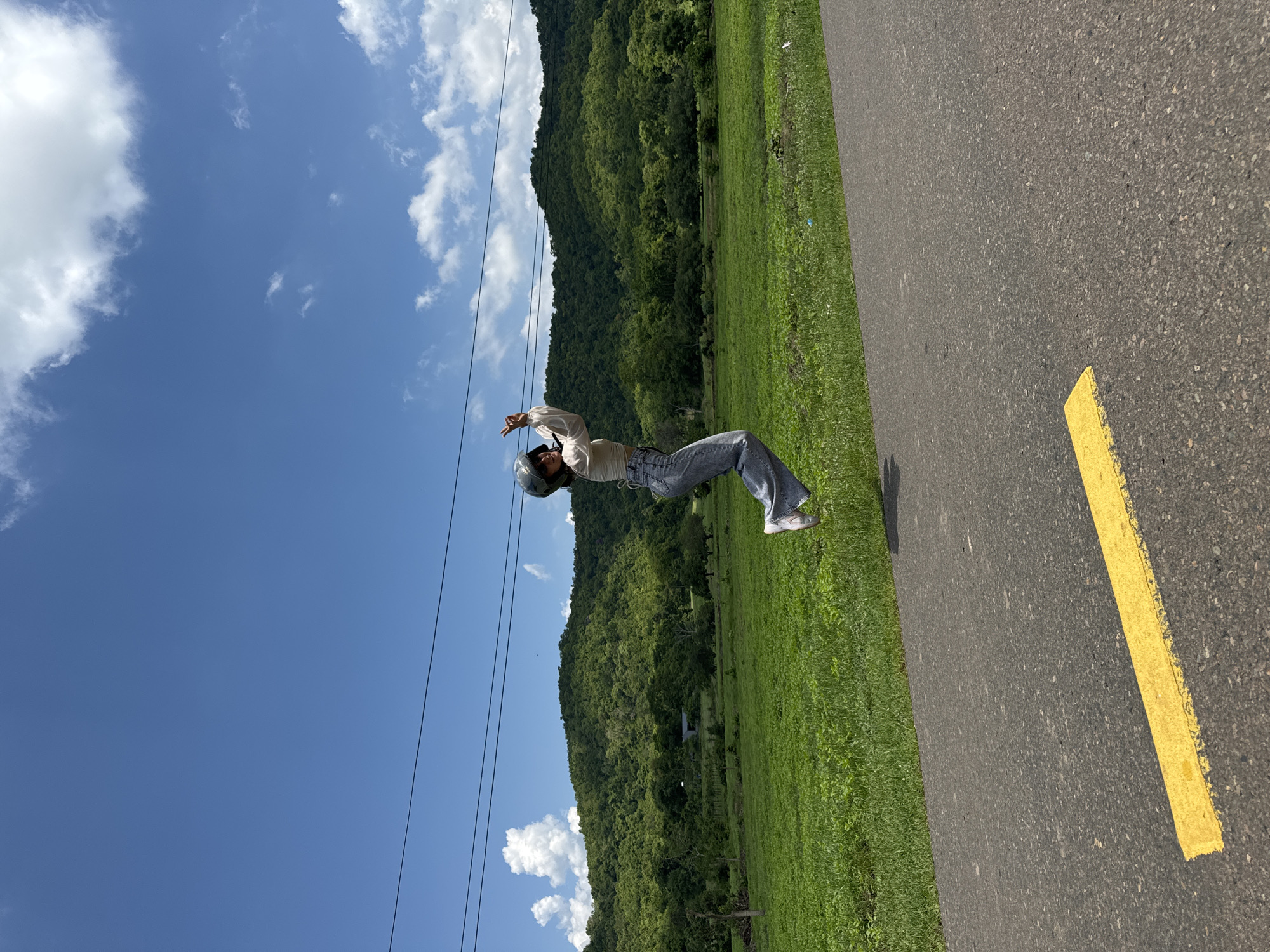 A person is captured mid-air, jumping on a paved road with a yellow line. They are wearing a light-colored shirt, blue jeans, and a helmet, with their hands raised in a celebratory gesture. The background features a lush green field, a dense forest covering rolling hills, and a bright blue sky with scattered white clouds. Power lines are visible stretching across the upper portion of the image. The scene appears to be in a rural or natural setting, possibly within the city of Ham Thuan Bac, Vietnam, given the landscape.