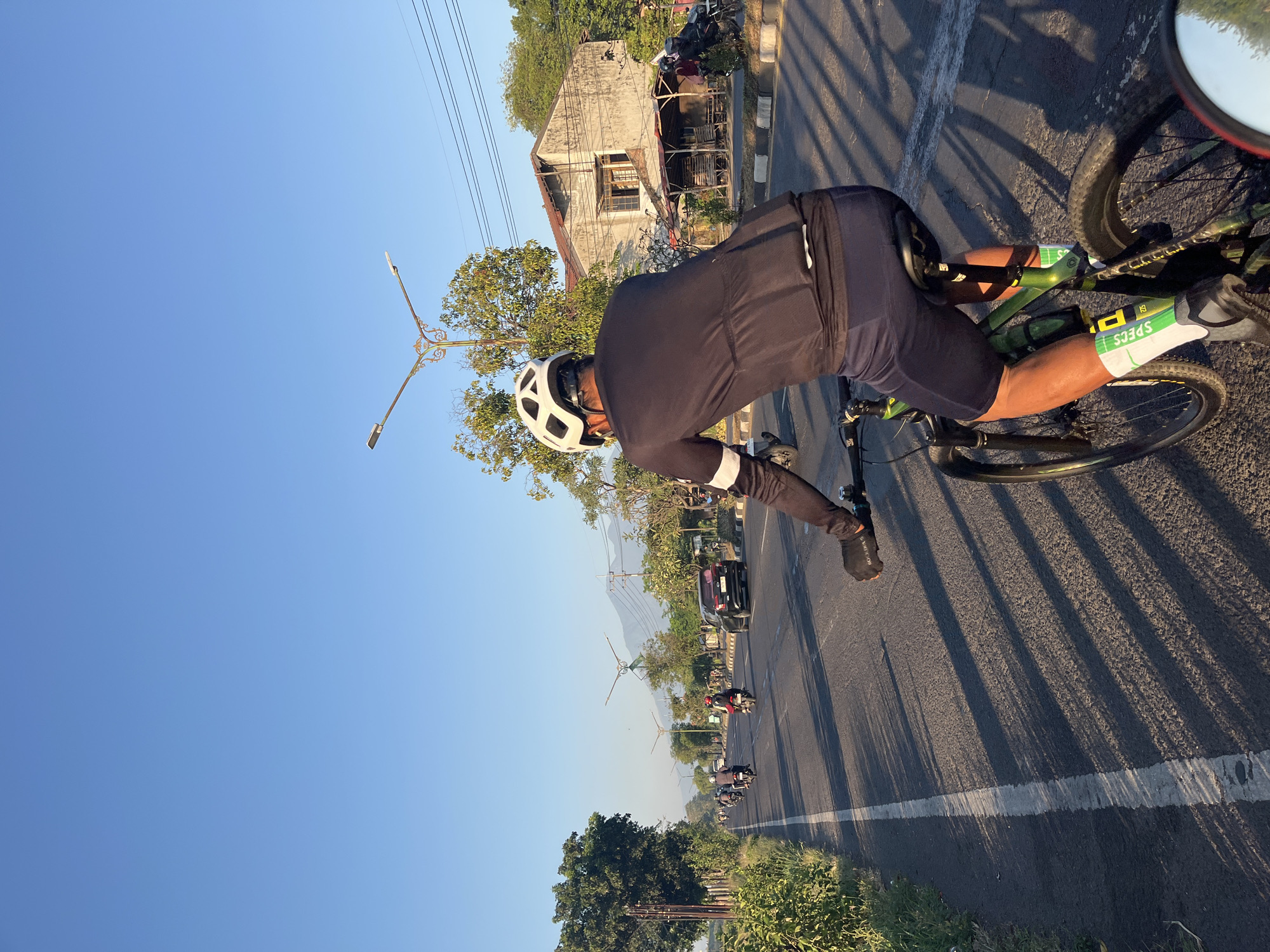 The image depicts a rear view of a cyclist wearing a white helmet, black long-sleeved cycling jersey, and dark shorts, riding a green mountain bike on an asphalt road. The cyclist's right hand, wearing a black glove with orange accents, is on the handlebar. The road is lined with a white dashed line and a solid white line. On the right side of the road, there is a worn, two-story building with a red tile roof and an adjacent structure with a thatched roof. Several motorcycles and a black car are visible on the road, suggesting moderate traffic. Lush green vegetation, including trees and bushes, borders the road. In the background, there are streetlights, power lines, and a distant mountain range under a clear blue sky. The scene suggests an outdoor activity in a semi-urban or rural environment, likely during the morning or late afternoon due to the long shadows cast by the cyclist and trees. The overall atmosphere is one of a sunny day with a relaxed pace of life.