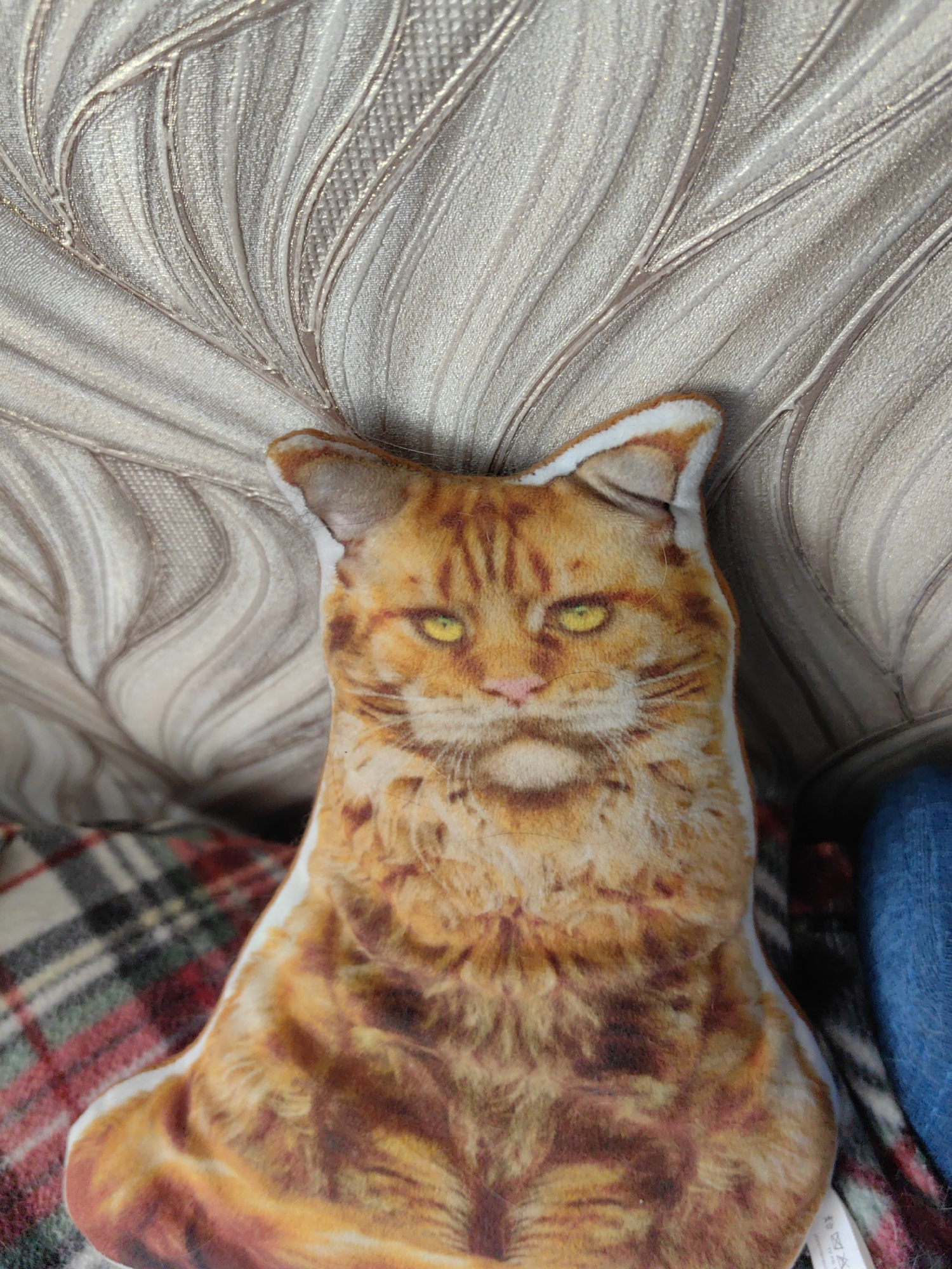 An indoor scene in the city of Unknown, Unknown, displays a plush, rectangular pillow designed to resemble an orange tabby cat. The cat pillow, featuring prominent yellow eyes, a pink nose, and distinct orange and brown fur patterns with white accents around the ears and facial outline, is positioned upright. Behind the pillow is a light beige or off-white textured surface, likely wallpaper or a headboard, embossed with large, stylized, undulating leaf or feather patterns that exhibit a subtle metallic sheen. Below and to the left of the pillow, a plaid fabric, possibly a blanket or throw, is visible, showing a tartan pattern in red, dark green, black, and white. To the right of the pillow, a partial view of a blue, textured object or fabric is present.