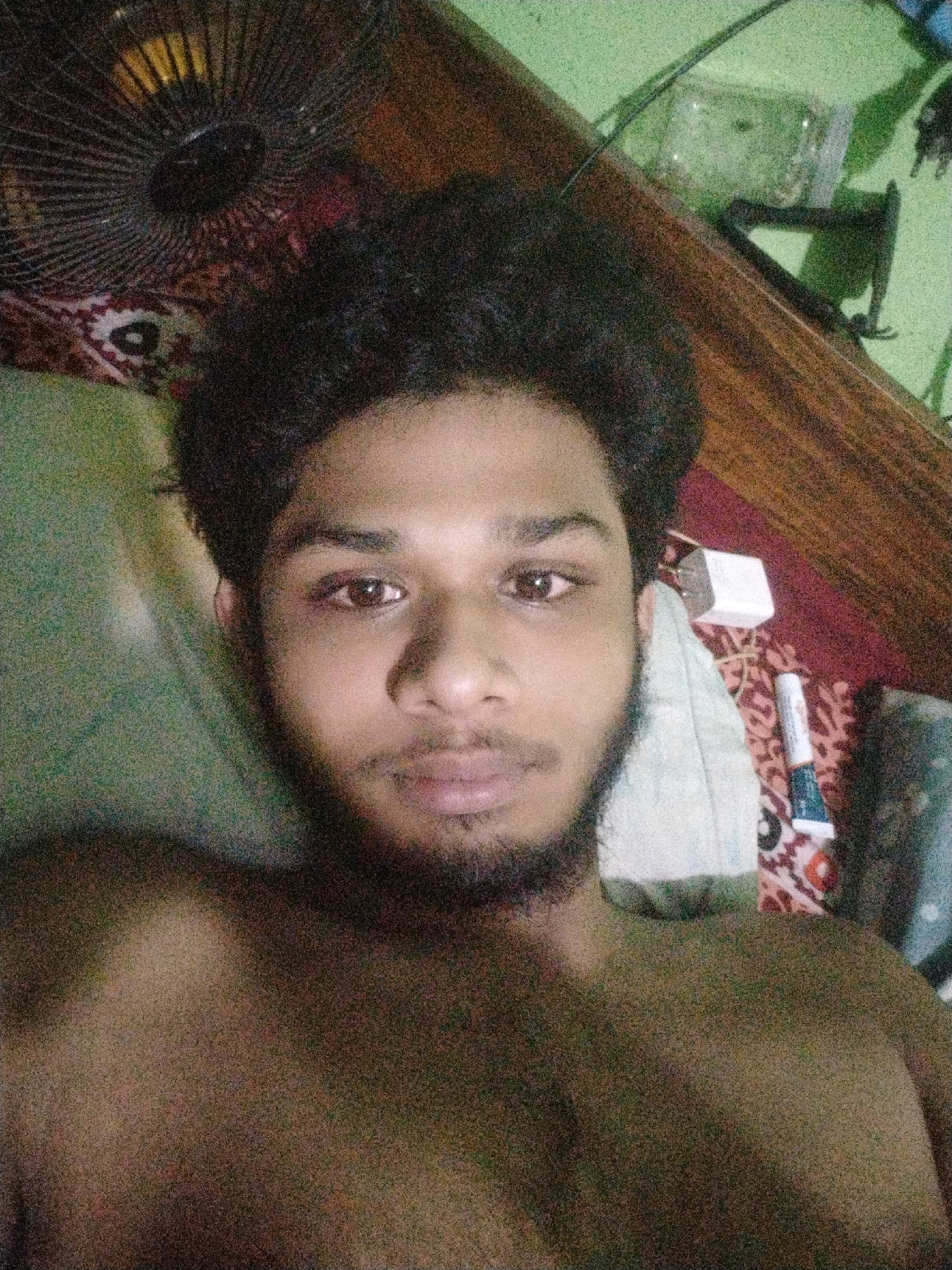 A selfie of a young man with dark, curly hair and a beard is taken from a low angle, showing his chest and upper body. He is lying down, with a fan visible in the background. The room appears to be a bedroom, with a wooden surface and a green wall. There are also some personal items on a patterned fabric, including a white box and a tube of what appears to be toothpaste or lip balm. The lighting is dim and grainy. The image was taken in Dhaka, Bangladesh.