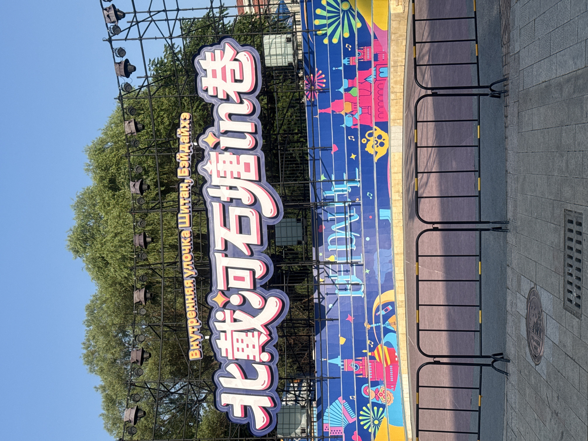 The image shows a large outdoor banner with Chinese text and colorful illustrations. The banner is supported by a black metal scaffolding structure, and several spotlights are attached to the top. The text on the banner appears to be in two languages: Russian at the top and Chinese below. The Chinese text, in large, stylized characters, reads "北戴河石塘in卷" (Běidàihé Shítáng in Juǎn). The Russian text above it reads "Внутренняя улочка Шитана, Байдайхэ" (Vnutrennyaya uločka Shitana, Baydajkhe), which translates to "Inner street of Shitan, Beidaihe".
The banner itself has a blue background and features a vibrant, cartoonish mural. This mural depicts various elements, including architectural structures resembling colorful castles or pagodas, musical instruments, an accordion, fireworks, and abstract shapes. The style suggests a festive or cultural event. Below the banner, black metal crowd control barriers with yellow reflective tape are erected, positioned on a paved area. In the foreground, there are patterned pavement stones and a manhole cover. The background consists of lush green trees under a clear blue sky.
This scene is located in Qinhuangdao, China, specifically in the Beidaihe district, which is known for its historical significance and tourist attractions. The reference to "Shitan" and "Beidaihe" on the banner confirms this location. The image captures a public space likely set up for an event or exhibition. No people are visible in the frame.