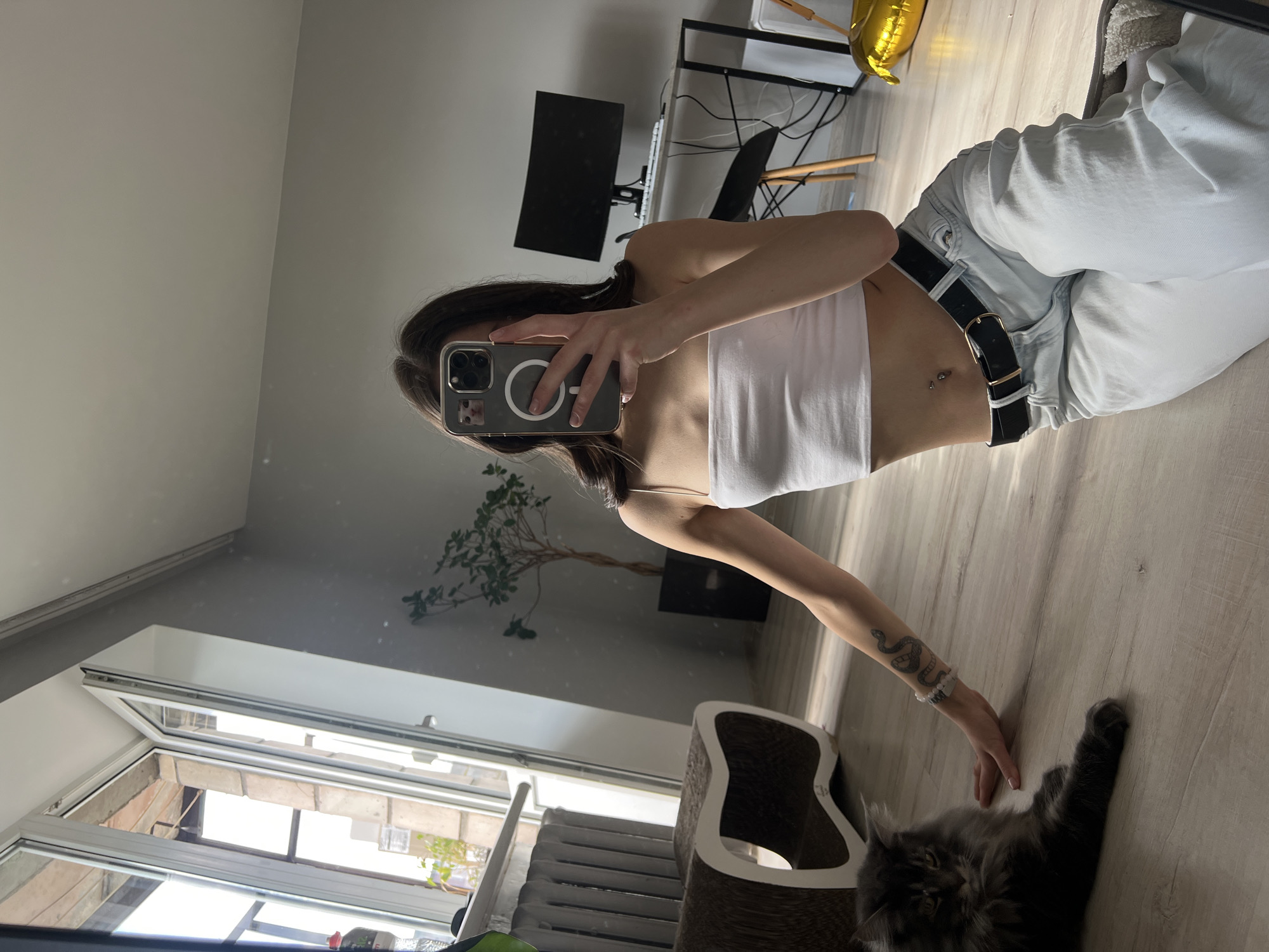 A young woman with long dark hair is sitting on the floor in a modern apartment, taking a selfie with her phone. She is wearing a white tube top, light blue jeans, and a black belt. Her belly button is pierced, and she has a snake tattoo on her left arm. A grey Maine Coon cat is lying on the floor next to her. In the background, there is a large window with a view of a building facade, a radiator, a scratching post for cats, a potted plant, a desk with a computer monitor, and a golden balloon. The floor is made of light-colored wood. The scene appears to be in Ternopil, Ukraine.