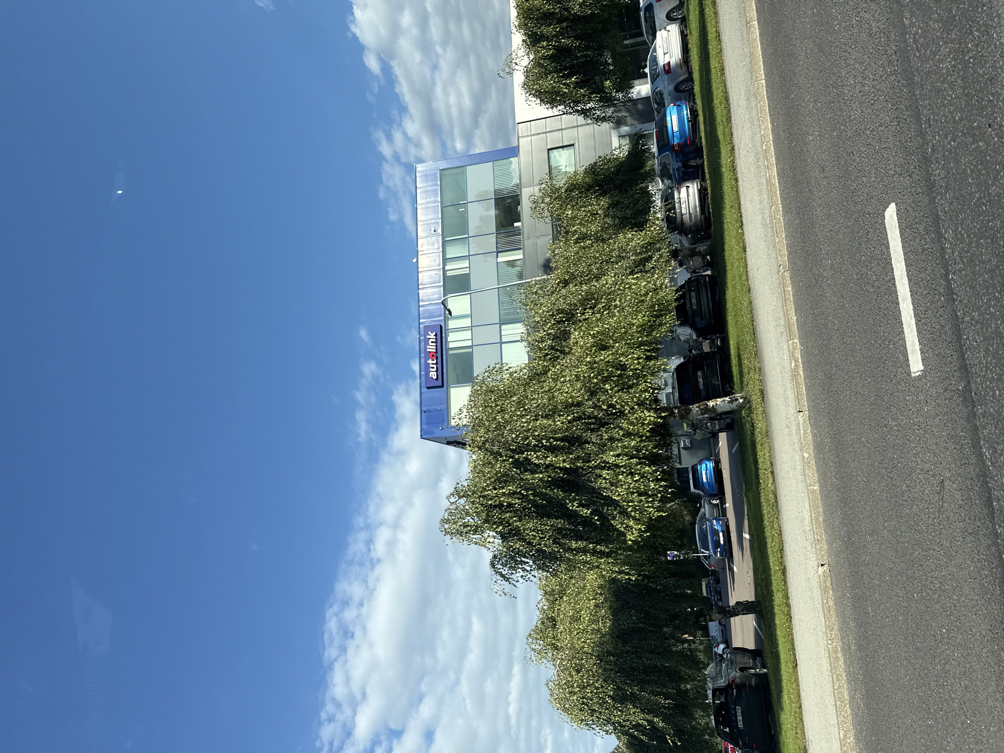The image depicts the exterior of a modern office building with the "autolink" logo visible on its facade, situated in Tallinn, Estonia. The building features a blue-tiled upper section with large glass windows and a gray lower section. In front of the building, a row of cars is parked on a grassy area, with more cars visible in the background. Several trees with dense, green foliage partially obscure the view of the cars and the building. The scene is set against a bright blue sky with scattered white clouds, indicating daylight. The foreground shows a portion of an asphalt road with a white line marking. There are no people visible in the image.