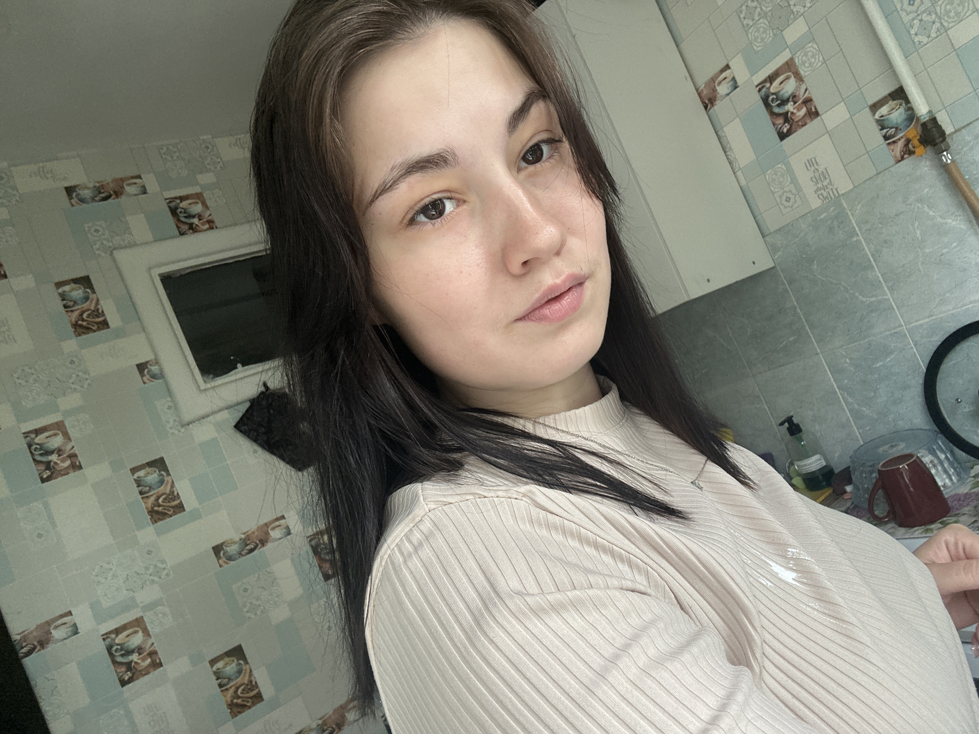 A selfie-style portrait features a young woman with long, dark brown hair and light skin looking directly at the camera. She wears a light-colored, ribbed, high-necked top, with a thin silver necklace visible around her neck. Her expression is neutral and direct.
The setting appears to be an indoor kitchen in Chaikovsky, Russia. Behind her, a tiled wall covers much of the background. The tiles are primarily light blue and grey squares with subtle patterns, interspersed with rectangular accent tiles. These accent tiles depict coffee cups, some with coffee beans and others with latte art. One of these tiles clearly displays the text "LIFE IS SHORT STAY SWEET" and another has "COFFEE". White or light grey kitchen cabinets are visible to the left and right sides of the frame. A window or dark opening is partially seen behind her left shoulder. To her right, a pump bottle (possibly soap), a dark red mug, and a pot lid or similar round object are on a counter. A light-colored pipe, possibly copper or brass, runs vertically along the wall. Natural light illuminates the scene, suggesting it is daytime.