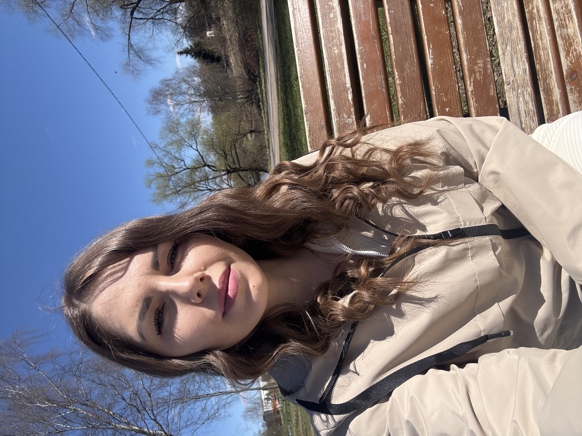 The image shows a young woman with long, brown, curly hair and light skin sitting on a wooden park bench. She is wearing a light beige jacket with a black zipper. The background shows a clear blue sky, trees with budding leaves, a green lawn, and a road. The location appears to be Shevlyakovo, Russia.