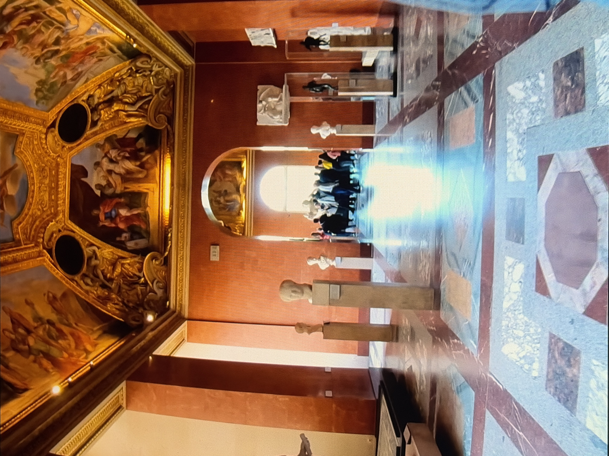 The image depicts the interior of a grand museum hall with high ceilings adorned with ornate golden frescoes depicting classical scenes. The walls are a rich, textured orange. The floor is a polished marble with a geometric pattern of red, blue, and cream-colored squares and octagons, reflecting the light from an unseen source and creating a shimmering effect.
In the foreground, several classical busts are displayed on tall, slender stone pedestals. The first pedestal, to the left, is empty or features a very faint object. The second pedestal holds a bust of a figure with a rounded head. The third pedestal displays a bust with more defined facial features. Further down the hall, another pedestal is visible, with a bust that appears to be partially obscured.
In the middle ground, a large arched opening leads to another section of the museum, where a crowd of people, mostly wearing casual clothing, are gathered and observing exhibits. They appear to be tourists or museum visitors. Beyond the archway, natural light streams in from large windows, illuminating the space.
Various sculptures are also visible on pedestals and display stands along the walls, including white marble sculptures and darker bronze figures. A small white rectangular sign is mounted on the wall to the right of the arched doorway.
The overall impression is one of historical grandeur and cultural richness, typical of a renowned art museum. The lighting suggests it is daytime.