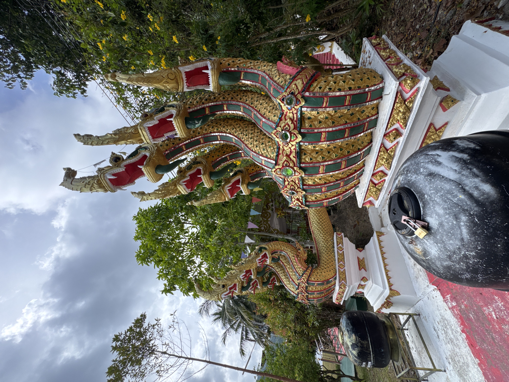 The image shows a low-angle, outdoor shot of a large, ornate statue of a multi-headed serpent, likely a Naga, in a lush, green environment. The statue is gold and red, with intricate scales and decorative elements. It is positioned on a white, tiered pedestal adorned with golden motifs. In the foreground, two large, black, cylindrical objects are visible. The one on the right has two padlocks on its lid. The background features dense foliage, trees, and a cloudy sky. The location is identified as Koh Phangan, Thailand, specifically Tambon Koh Phangan.