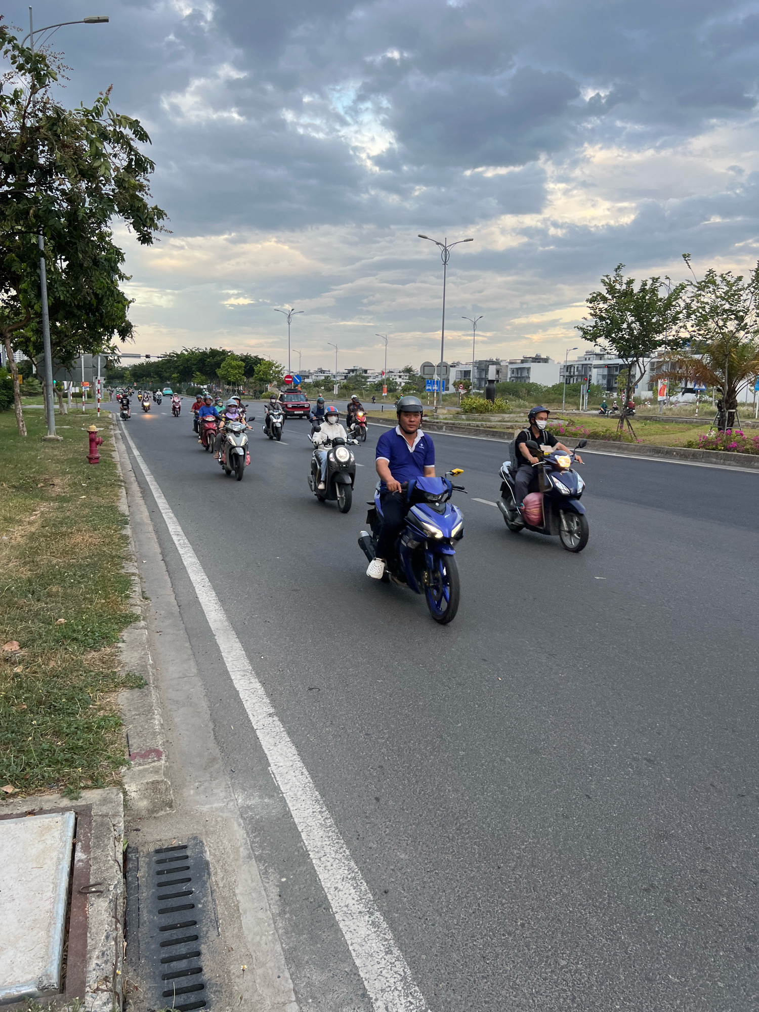 A long shot from a slightly elevated perspective shows a road with multiple lanes in Nha Trang, Vietnam. A line of motorcyclists, some wearing helmets and masks, are traveling in the same direction. The sky is overcast with a mix of gray and white clouds. Trees and buildings are visible on either side of the road, with some of the buildings appearing to be under construction. The road itself is paved and has white lane markers. A red fire hydrant is visible on the grassy median strip to the left of the road, along with a storm drain on the edge of the road. The overall impression is of a typical street scene in an urban or suburban area.