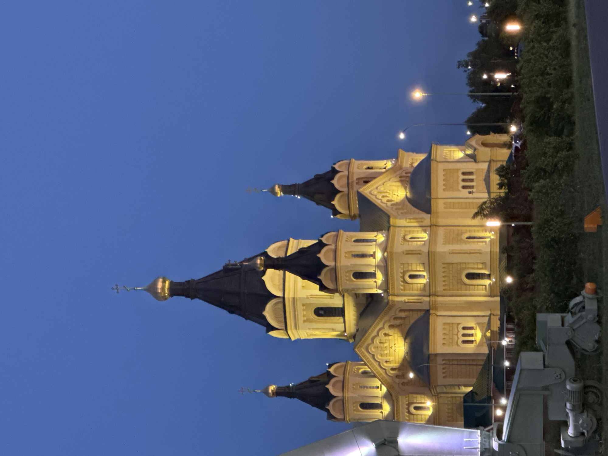 The image shows a church with multiple domes, illuminated by warm lights against a deep blue twilight sky. The architecture appears ornate, with decorative carvings and arched windows. Several onion domes, topped with crosses, rise from the building. The image is oriented vertically, with the church dominating the center.
To the right of the church, there is a line of trees and foliage, also illuminated by scattered lights from street lamps or possibly string lights. In the foreground and extending to the right edge of the frame, there are some industrial-looking metal structures, appearing to be part of some kind of machinery or equipment. One such structure has a cylindrical component that resembles a motor or pump.
The time of day is clearly evening or twilight, given the dark sky and artificial lighting. The weather appears clear. The location is identified as Nizhny Novgorod, Russia, which aligns with the architectural style of the church. There is no visible text in the image. The scene evokes a sense of grandeur and peacefulness, characteristic of illuminated religious architecture at dusk.