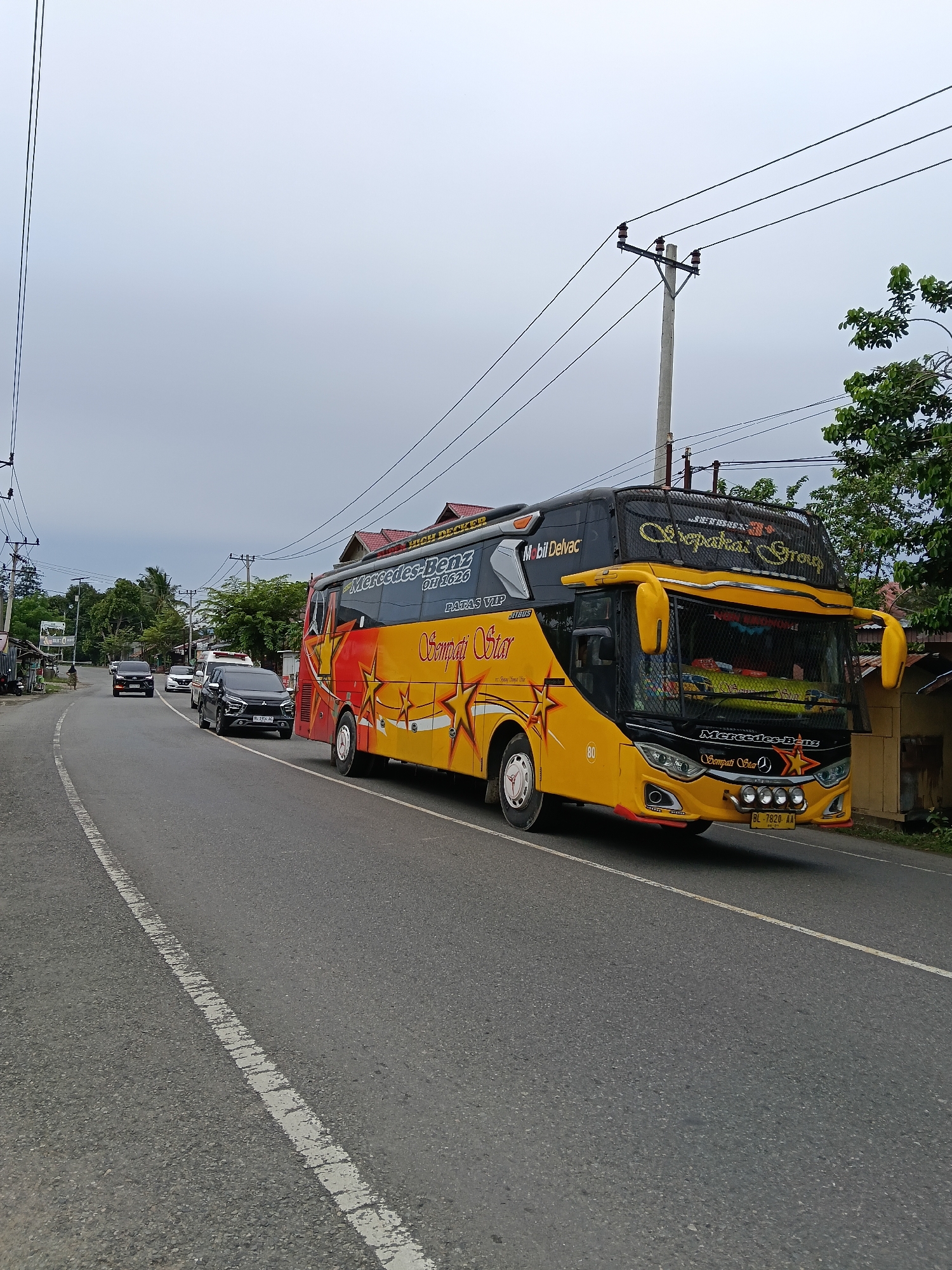 This image captures a bright yellow bus with red and gold accents driving down a paved road in Kuta Pungwa, Indonesia. The bus is branded with "Mercedes-Benz" and "Sempati Star." The road is lined with trees and utility poles, and a few other vehicles, including cars and a smaller van, are visible on the road ahead of the bus. The sky is overcast. The scene appears to be a typical day in this Indonesian town.