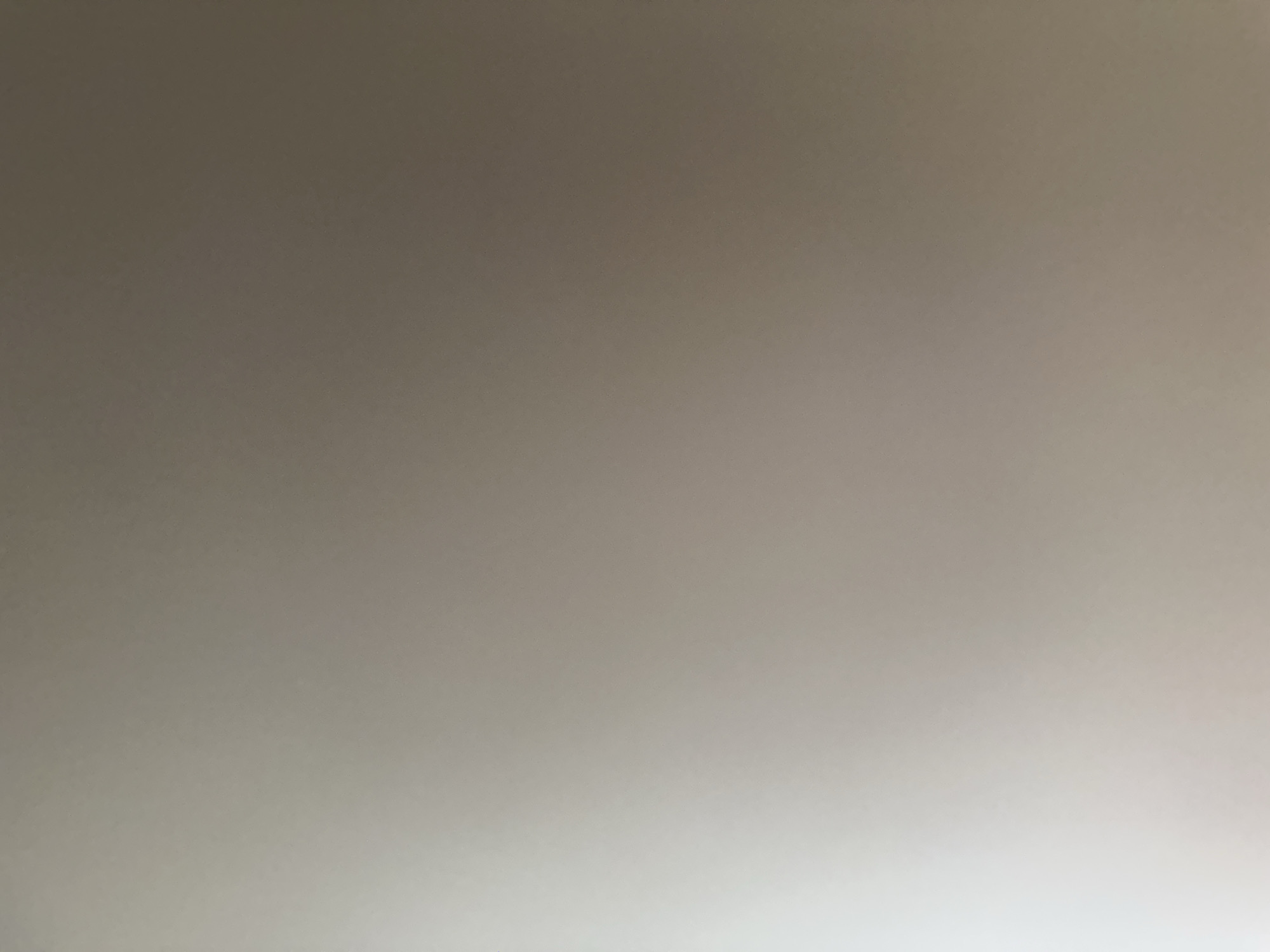 The image is a very blurry, close-up shot of a wall with a subtle gradient of light. The color ranges from a muted gray in the upper left to a brighter, off-white in the lower right. There are no discernible subjects, objects, text, or any indication of an activity or event. The lighting suggests a bright light source is present, possibly a window or lamp, illuminating the wall from the right and below. Given the location context of Belgrade, Serbia, it is likely an indoor scene, potentially a residential or office space. No specific time of day, weather conditions, or emotions can be inferred.