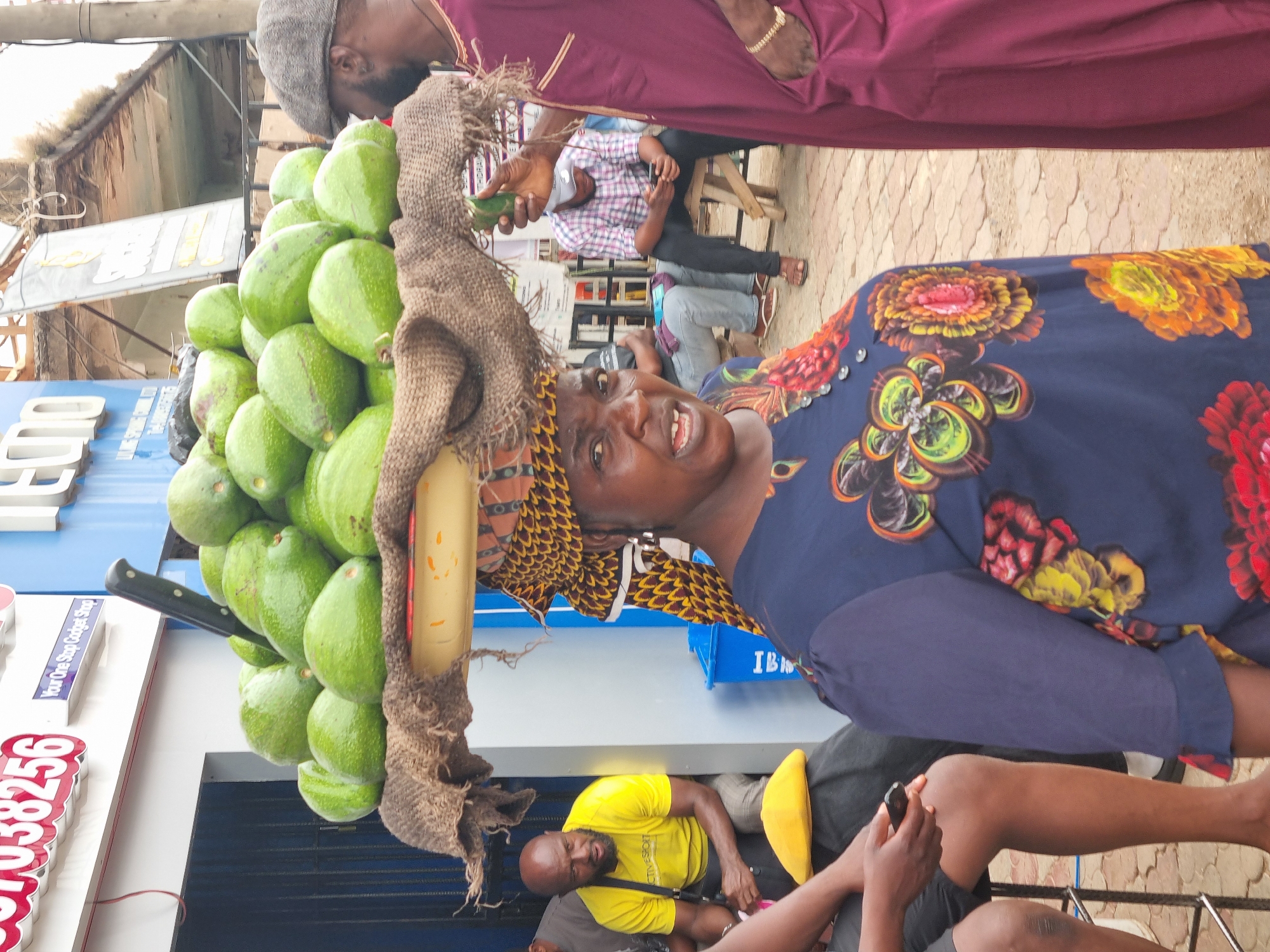 This image captures a vibrant street scene, likely in an urban or market area. The central subject is a woman carrying a substantial pile of green avocados balanced on her head. She wears a dark blue top adorned with large, colorful floral patterns and a patterned headscarf. She looks directly at the camera with an engaging, friendly expression, her mouth slightly open as if speaking or smiling. A piece of burlap or rough fabric is used to help secure the large load, and a dark-handled knife is visibly tucked into the avocados, suggesting they might be prepared for sale.
In the background, several other individuals are visible. To the left, a man in a grey cap and purple/maroon attire is partially seen. Further back, another man in a plaid shirt and dark trousers sits on a low stool, appearing to be engrossed with a mobile phone. In the lower foreground, a man wearing a bright yellow t-shirt and dark pants is seated, also looking towards the camera. Other indistinct figures are present in the bustling scene.
The setting includes various buildings and structures, indicative of a commercial street. Visible text on signs includes "008", "Your One Stop Gadget Shop" on a blue sign, and a partial phone number "07038256" in red on white. The ground appears to be paved. The bright lighting suggests it is daytime, and the overall atmosphere is one of daily commerce and interaction in Jos, Nigeria.