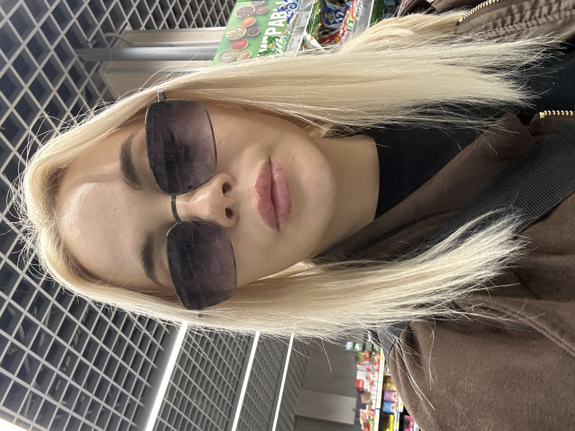 A woman with blonde hair and sunglasses stands in an indoor location. Behind her is a wall with a grid pattern and shelving filled with various products, possibly in a store or market in Megion, Russia. She is wearing a dark shirt and a brown jacket with a strap across her shoulder.
