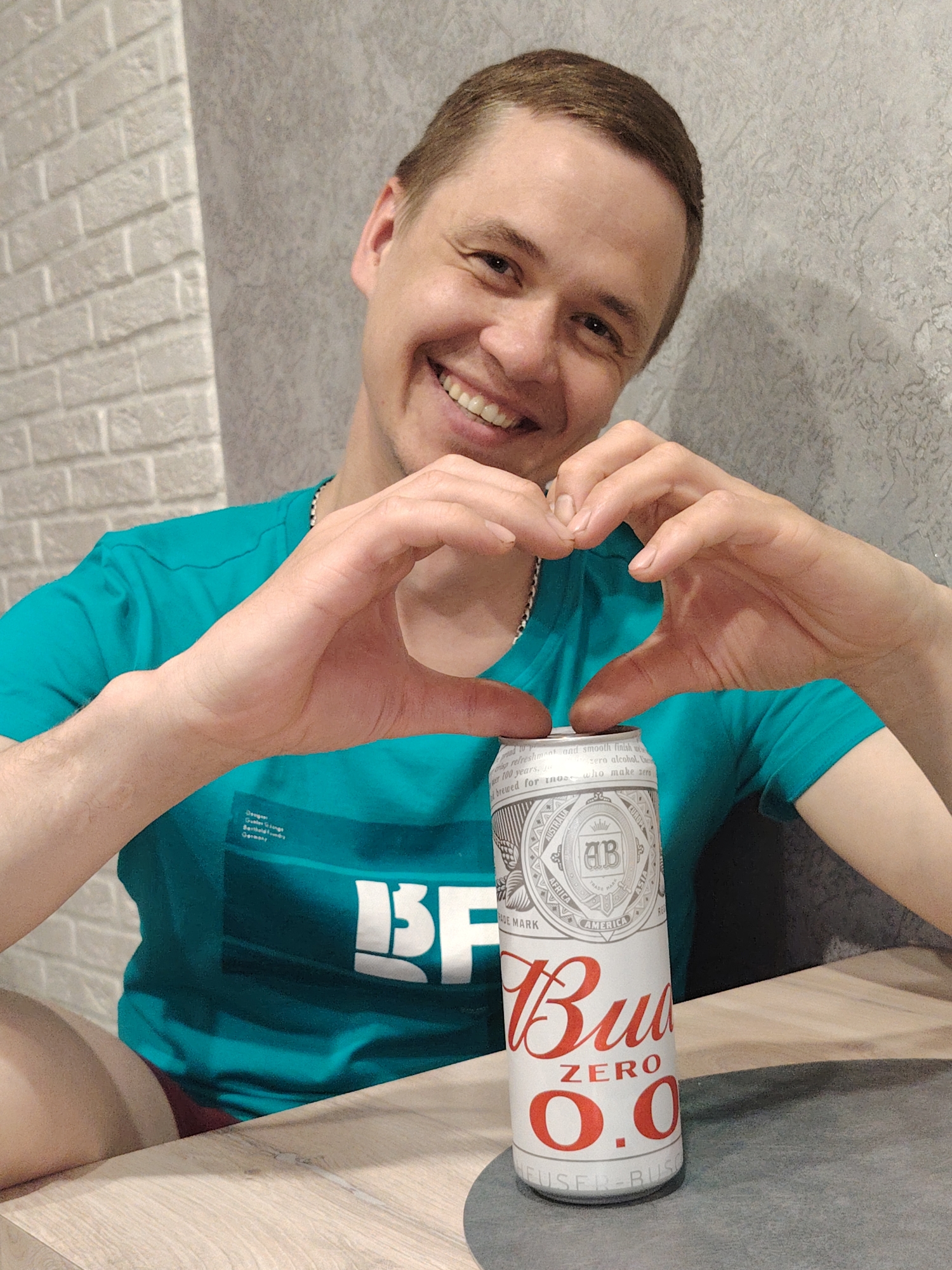 A cheerful man, likely in his 20s or 30s, is featured in a close-up, seated at a light wooden table indoors. He is smiling broadly with a bright, open expression, looking directly at the viewer. His hands are positioned to form a heart shape, encompassing the top of a white can of "Bud Zero 0.0" non-alcoholic beer. His thumbs meet over the can's opening, reinforcing the heartfelt gesture.
The man has short, neatly styled brown hair and fair skin. He is wearing a teal or turquoise t-shirt with visible text on the chest, including "Designer Gunter Grings Bertholdo Germany" and large white letters that appear to be "ISF" and "BF". A thin metal chain is visible around his neck.
The can of "Bud Zero 0.0" is the central object, prominently displaying its branding in red text. Additional text on the can includes the "AB" logo, "TRADE MARK", and mentions of various continents like "AUSTRALIA", "AFRICA", "AMERICA", and "ASIA". Descriptive phrases such as "refreshment and smooth finish" and "zero alcohol" are also legible, along with the slogan "brewed for those who make zero".
The setting appears to be a modern indoor space, possibly a home or casual establishment in Chaikovsky, Russia. The background features a light grey brick wall on the left, contrasting with a darker, textured grey wall on the right. The lighting is soft and even, highlighting the man's joyful expression and the product. The overall scene conveys a feeling of happiness and positive endorsement for the beverage.