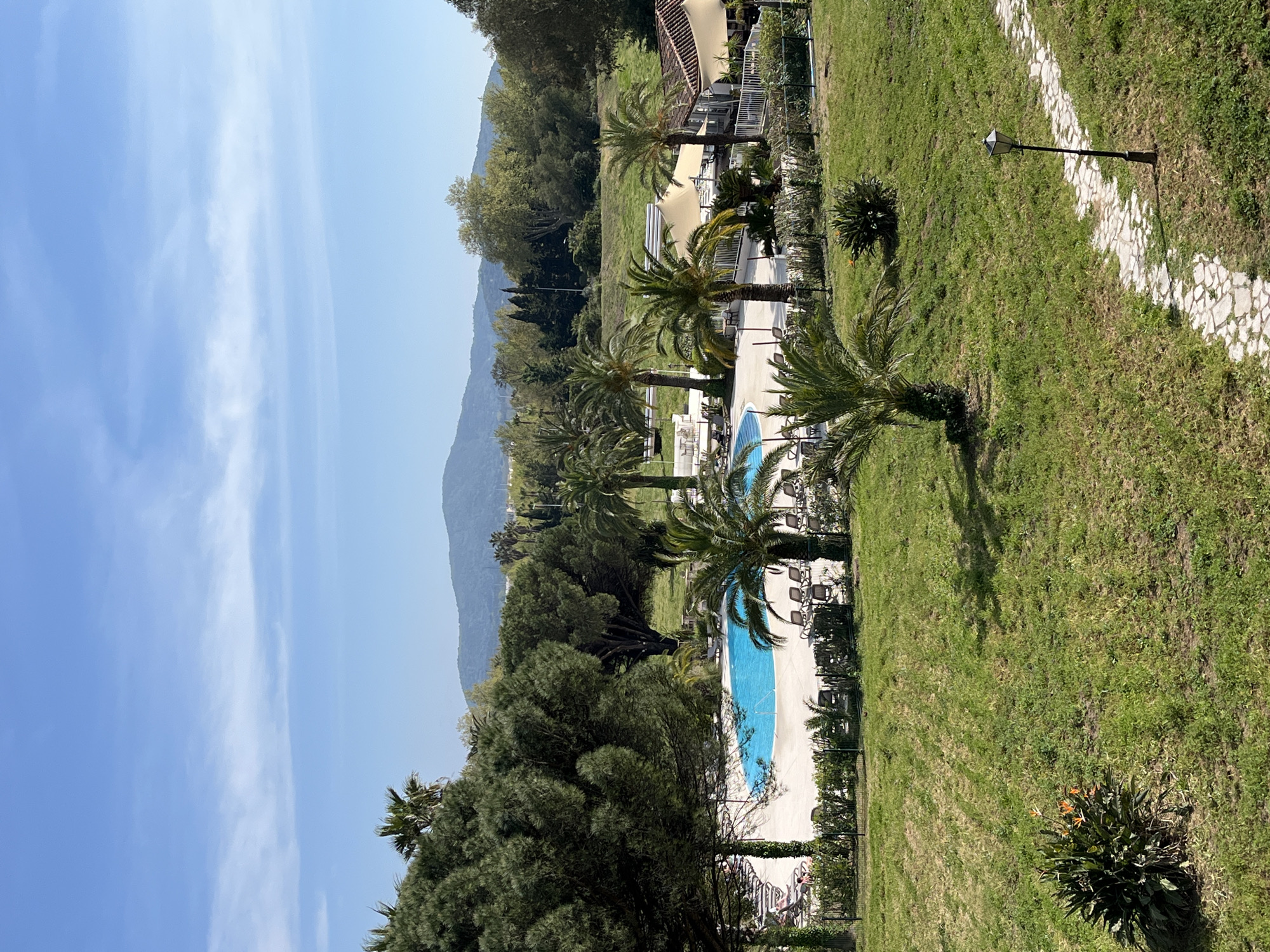 The image shows a landscape in Grimaud, France, featuring a swimming pool area and surrounding natural elements. The scene is captured from an elevated perspective, possibly from a higher floor of a building.
The sky above is mostly clear with a few elongated white clouds. In the background, a mountain range provides a natural backdrop. The middle ground features a resort-style area with two swimming pools with blue water, each bordered by white tiling and lounge chairs. Palm trees are scattered throughout the pool area, providing shade and adding to the tropical ambiance. Manicured green lawns border the pool area. A stone path runs along the edge of the lawn, with a lamppost situated beside it.
In the immediate foreground, there's lush greenery with trees and bushes. A bush with orange flowers is visible near the bottom right corner.