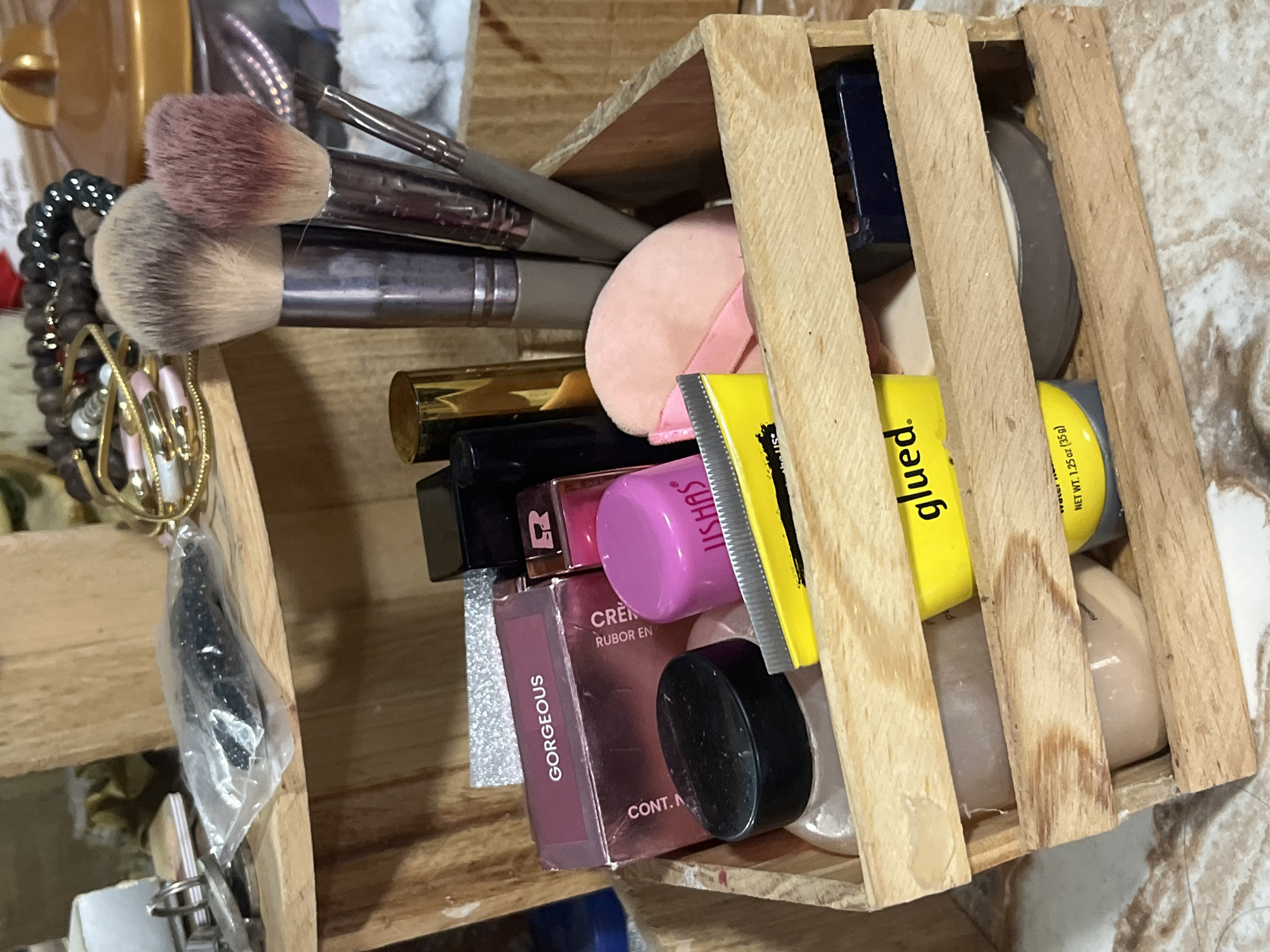 A collection of beauty and personal care items is neatly organized within a rustic wooden crate. The subjects include several makeup brushes with varied bristle colors (brown, beige, pink-tinged), along with jewelry such as beaded bracelets and a necklace featuring a black charm encased in plastic.
Various cosmetic products are visible, including a soft pink makeup sponge, lipstick tubes in gold and black, a small black-lidded jar, and a clear bottle containing a white liquid. Also present are a blue compact, another compact with a light brown/beige pan, and a silver glittery item, possibly nail polish.
A prominent yellow tube labeled "glued." is seen, along with a pink-capped product from the brand "USHAS". A rectangular box, likely for a cream blush, displays the text "GORGEOUS", "RUBOR EN CRÈM" (Spanish for 'cream blush'), "CONT.", and the letter "R". The "glued." tube also states "NET WT. 1.25 oz (35g)".
The items are stored at an angle within the wooden crate, which has slatted sides. The background shows a light-colored surface, possibly a vanity or table, with a faint patterned texture. The scene depicts organized personal effects, likely in a home setting in Juan Griego, Venezuela. No specific activity is occurring, and the time of day is not discernible.