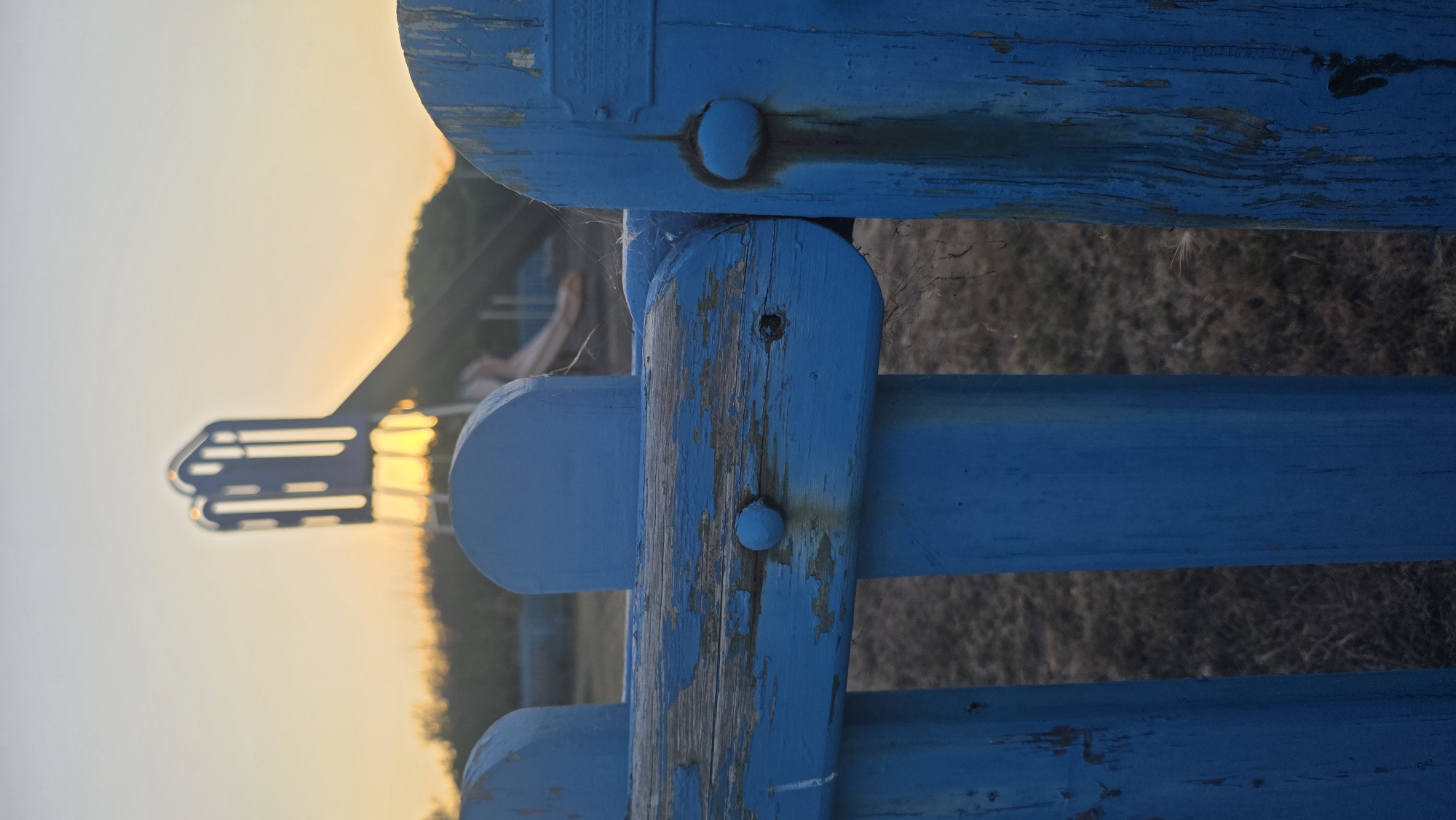 The image shows a close-up view of a weathered blue wooden fence in Rother, United Kingdom. The fence has peeling paint, revealing aged wood underneath, and some rust stains are visible on the hardware. In the background, out of focus, a children's playground slide is visible against a sunset sky. The ground is covered with dry grass. The scene is set outdoors, suggesting a park or recreational area.