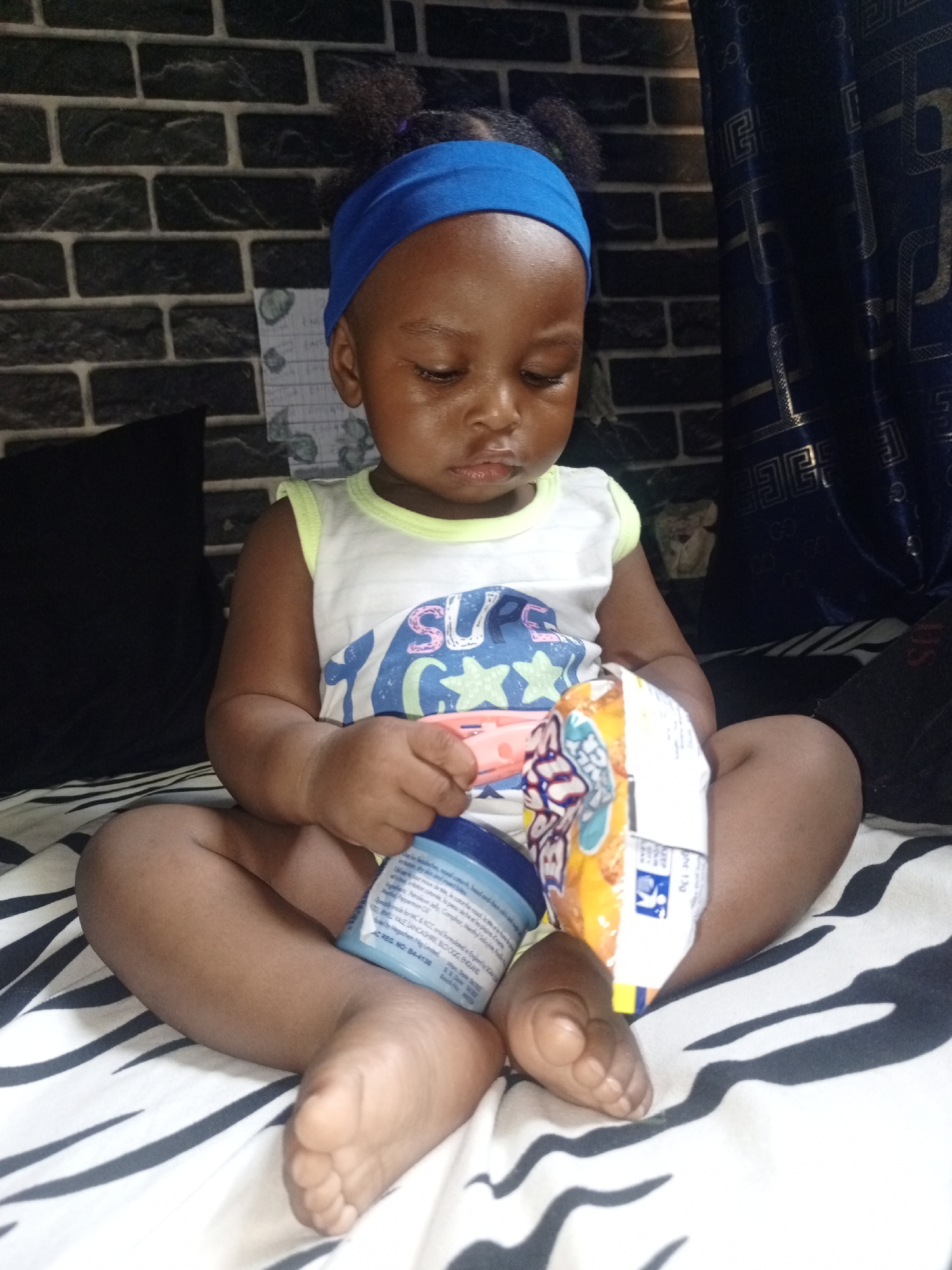 A young child, likely a baby or toddler, is seated cross-legged on a bed or soft surface, looking downwards with a focused expression. The child has dark skin and is wearing a white sleeveless top with a partial design that reads "SUPE" in blue and other colors. A bright blue fabric headband adorns their head, and their dark hair is styled in small puffs.
The child holds two items: a light blue plastic container in their left hand, and a white and colorful snack bag in their right hand. The snack bag features numbers like "49", "88", "130", and the word "GO", along with star graphics. The light blue container has several lines of white text, including "AC REG. NO: 84-4138", "NEL VALE LANCASHIRE, SUOOOG GANG", and "Some for MC & KCC underw".
The setting is indoors. The child is sitting on a surface covered with a distinctive black and white striped sheet, resembling a zebra pattern. To the child's left, a black pillow is visible. The background consists of a dark, brick-patterned wall. To the right, a dark blue curtain with a golden, intricate pattern is partially drawn. The lighting suggests an indoor environment, but the time of day is not discernible. The child appears to be engrossed in examining the objects in their hands.