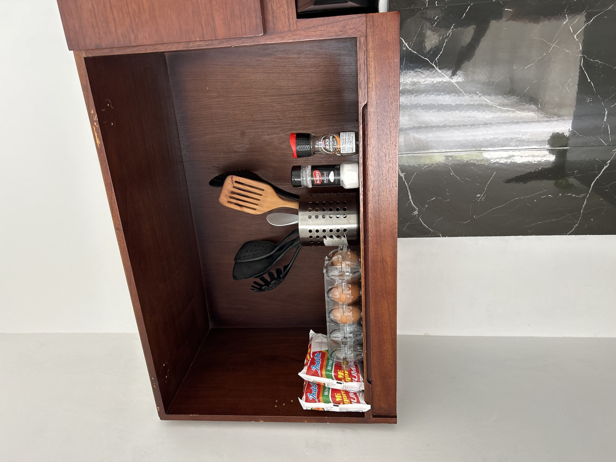 This is a medium-angle, eye-level, full shot of a dark brown, wooden kitchen cabinet shelf in Tembuku, Indonesia. The shelf is filled with various kitchen items. On the left side of the shelf are two packages of Indomie Mi Goreng instant noodles. To the right of the noodles is a clear plastic carton containing four brown eggs. In the center of the shelf is a stainless steel utensil holder containing several kitchen utensils, including a wooden spatula and a black slotted spoon. To the right of the utensil holder are two spice grinders: a white one labeled "La Fancy Sea Salt" and a black one labeled "Black Peppercorns." The shelf is mounted on a white wall, and below the shelf is a black and white marbled countertop or backsplash. The overall lighting is bright and natural, with shadows cast on the wall and shelf.