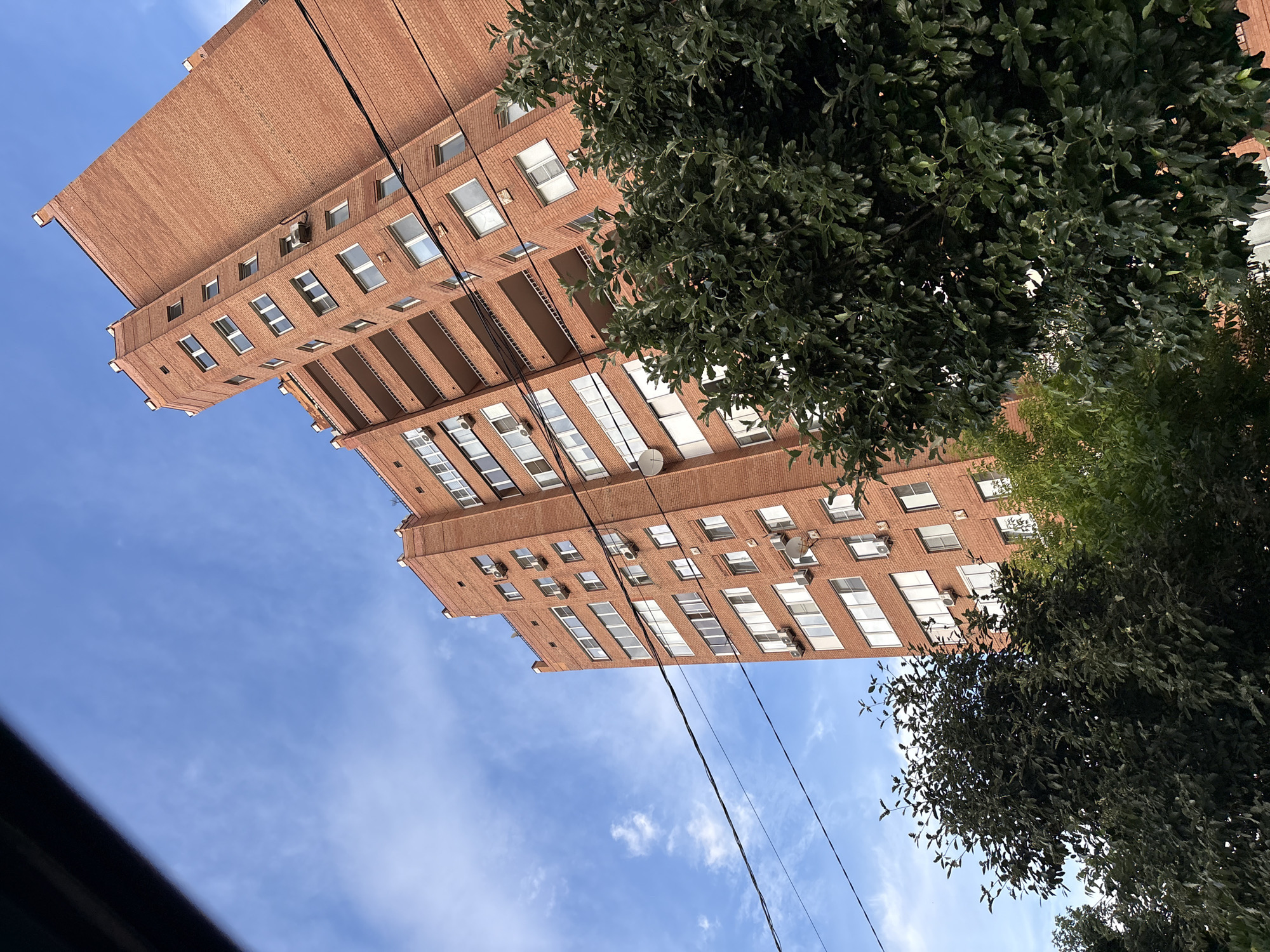This media file is a photograph taken outdoors, looking up at a tall, multi-story brick building. The building appears to be residential, with numerous windows visible on its facade. Some of the windows are open, and several air conditioning units are mounted on the exterior walls. The architecture suggests it is an older building.
Adjacent to the building, and partially obscuring it, are dense green trees with many leaves. The sky is visible behind the building and trees, appearing blue with scattered white clouds, indicating daytime. The sun is not directly visible, but the lighting suggests it is a bright day, possibly late morning or early afternoon.
Several black electrical wires run diagonally across the frame, suspended in front of the building and sky. These wires, along with the style of the building and the presence of air conditioning units, might suggest an urban or suburban setting.
The image is oriented with the top of the frame appearing towards the bottom of the scene as if the viewer is looking upwards. There is no text visible in the image. The overall impression is one of a typical day in a developed area, focusing on architectural and natural elements.